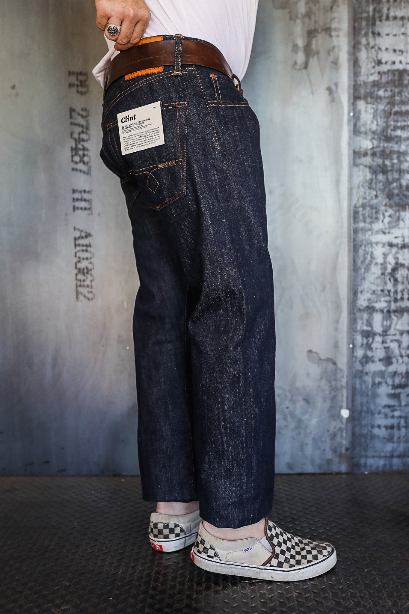 Person wearing dark raw denim jeans with a back pocket label and checkered Vans slip-on shoes