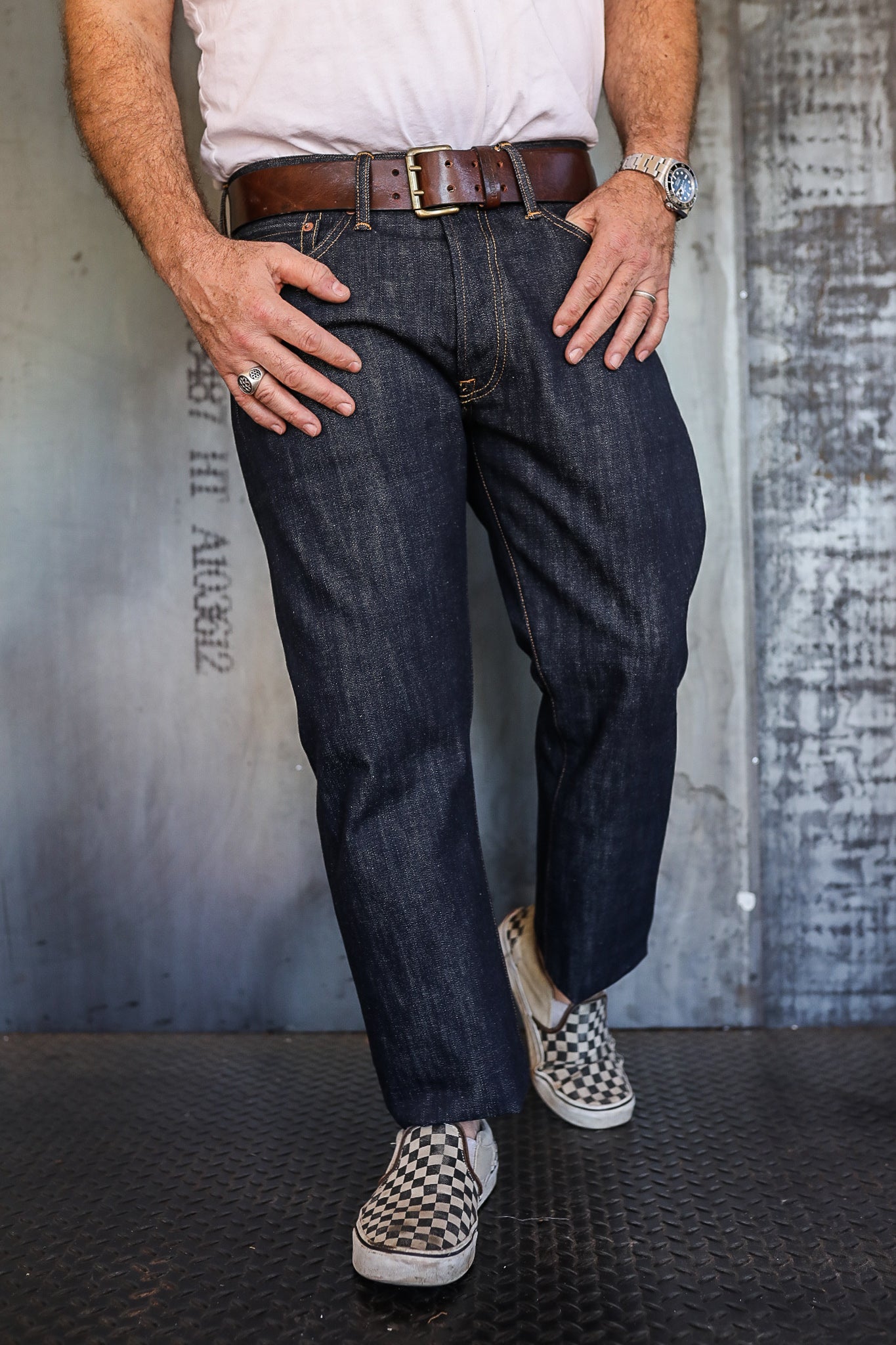 Man wearing dark raw denim jeans with brown leather belt and checkered slip-on shoes on metal floor