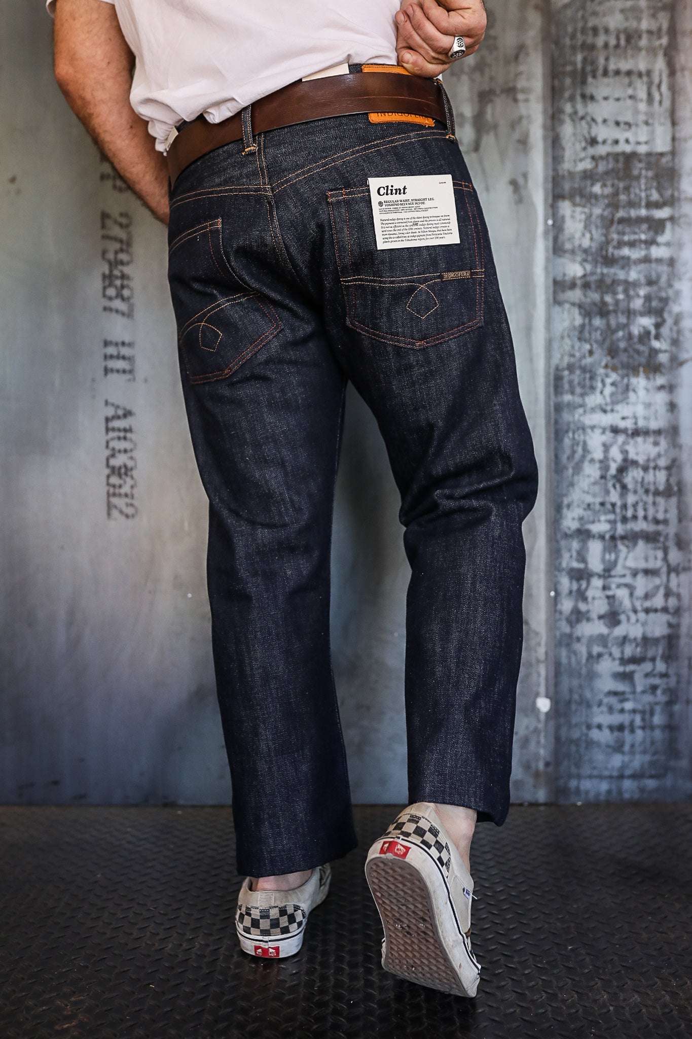 Rear view of man wearing dark raw denim Clint jeans and brown leather belt with checkered slip-on Vans shoes