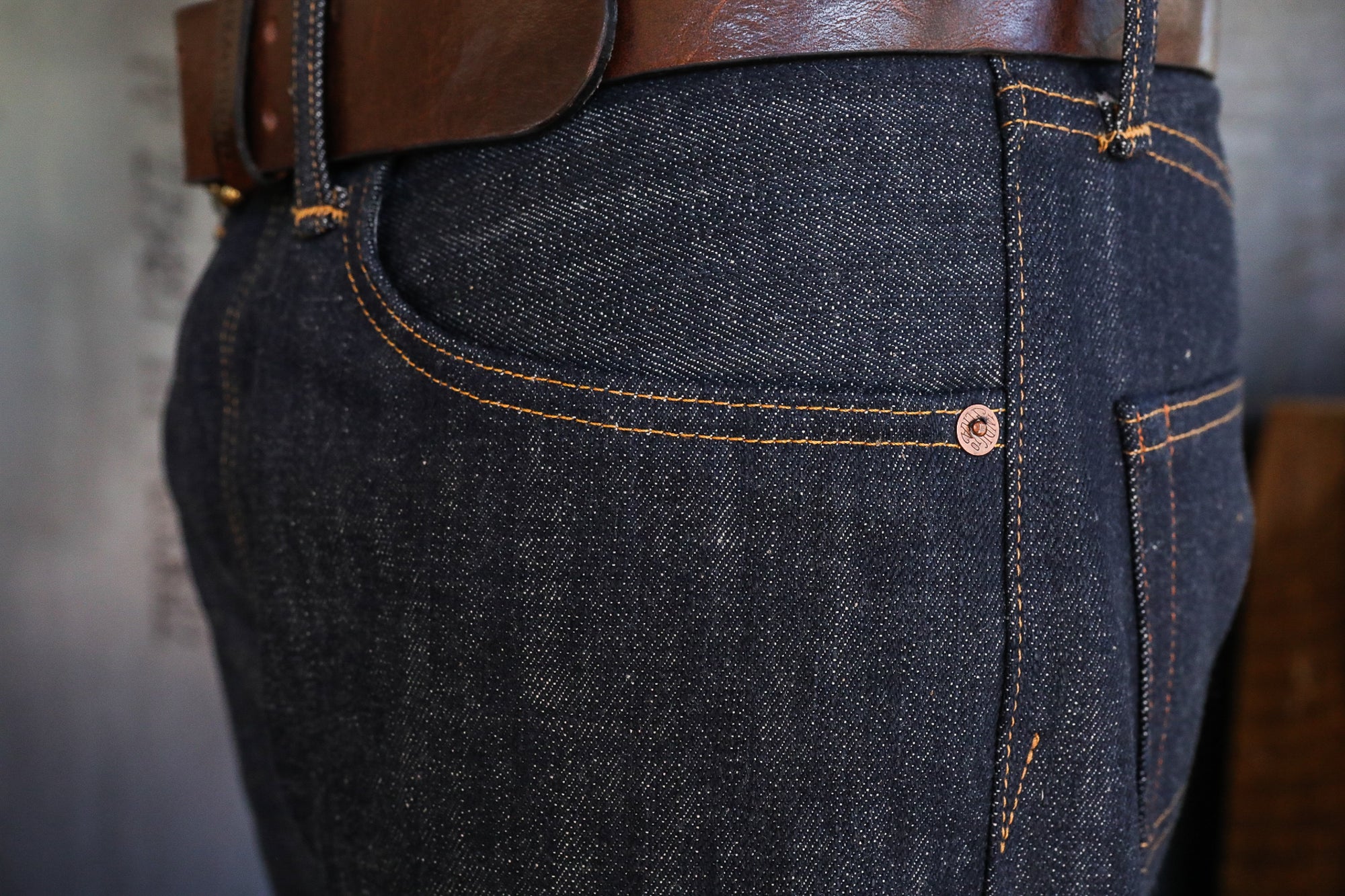 Close-up of dark raw denim jeans pocket with contrast orange stitching and brown leather belt