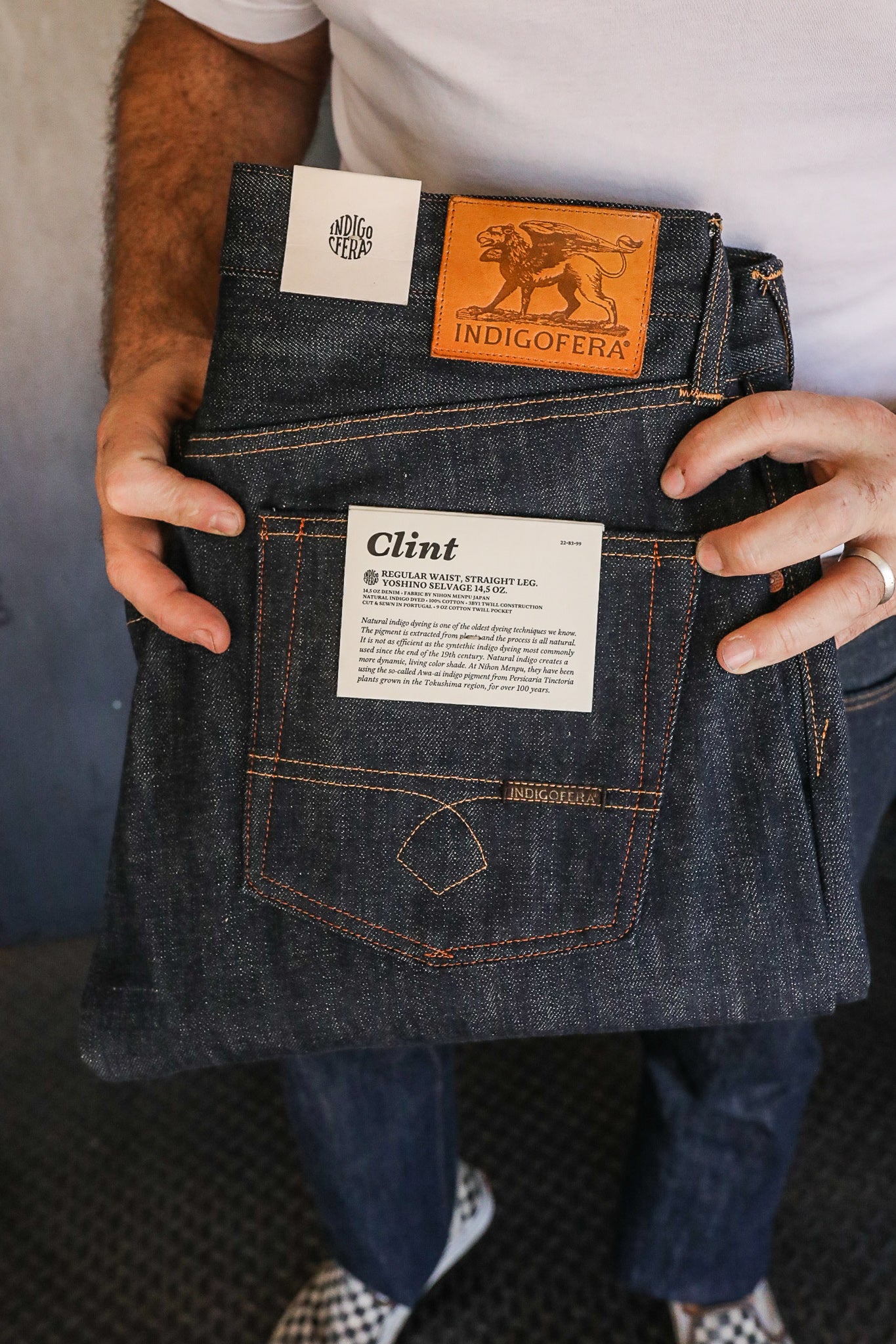Hands holding folded Indigofera Clint regular waist straight leg raw denim jeans with leather logo patch and informative label