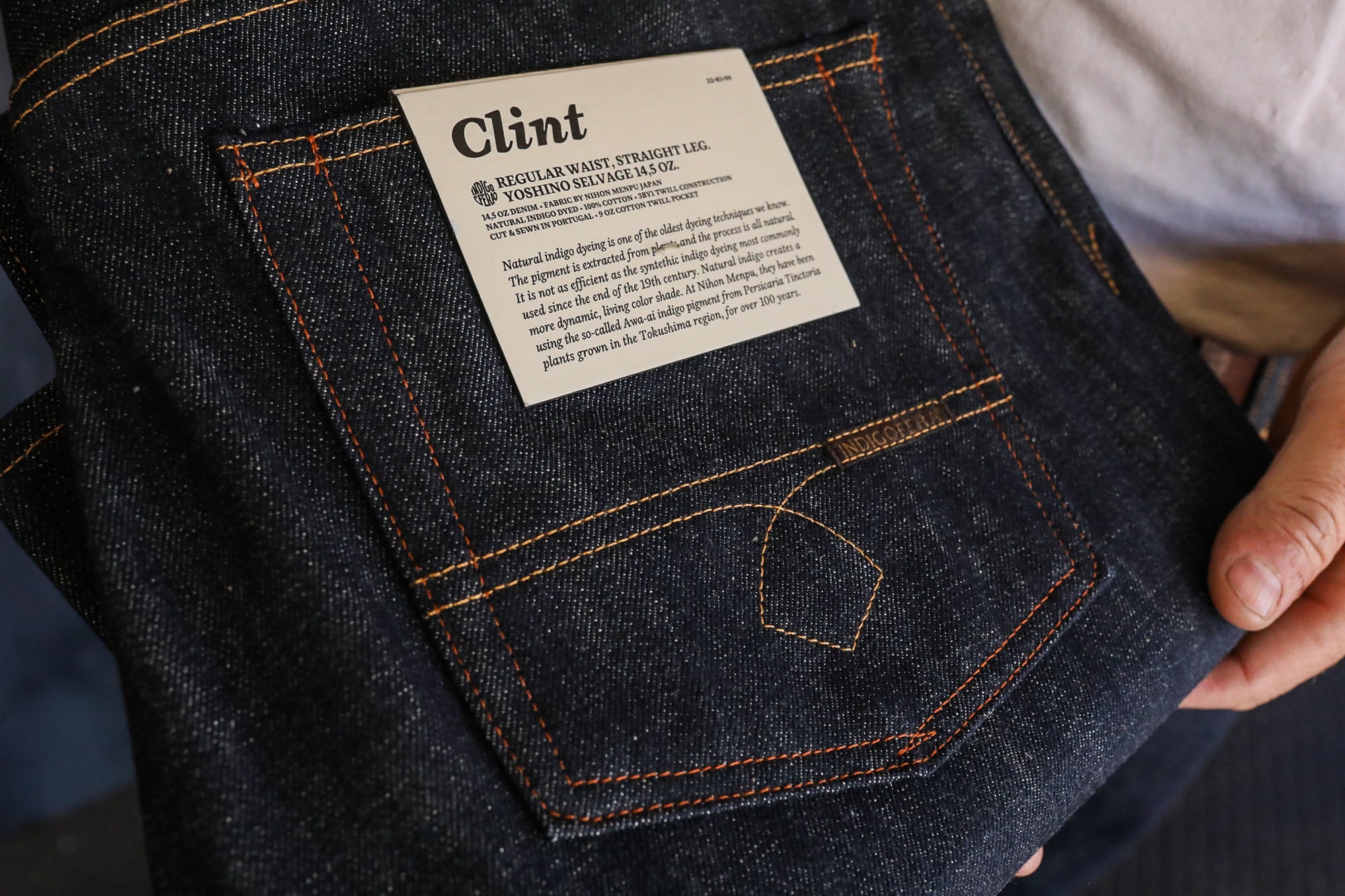 Hand holding dark indigo denim jeans with orange stitching and a 'Clint' label detailing natural indigo dye and fabric origin