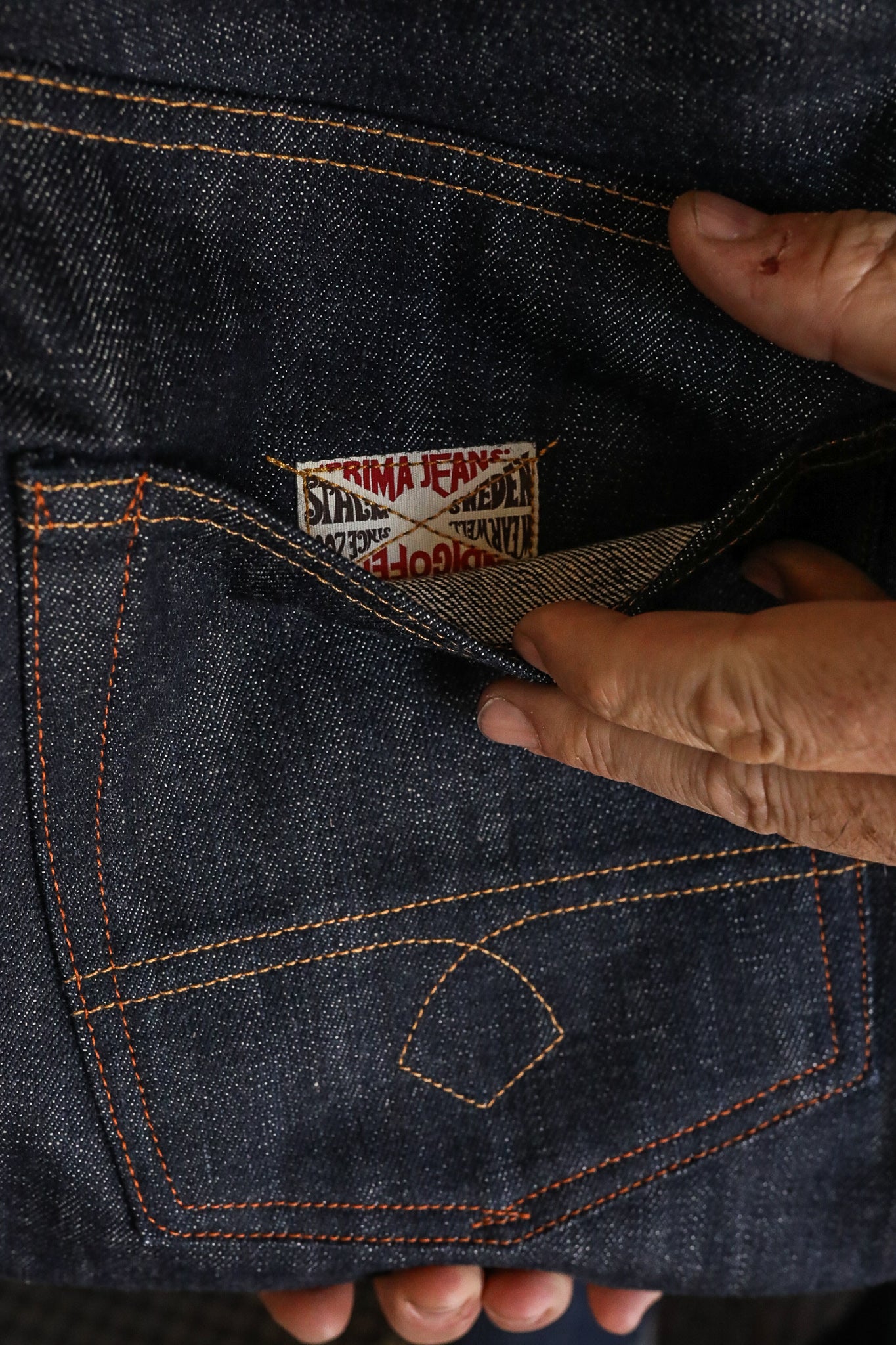 Close-up of hands holding dark denim jeans pocket with visible orange stitching and Prima Jeans label inside