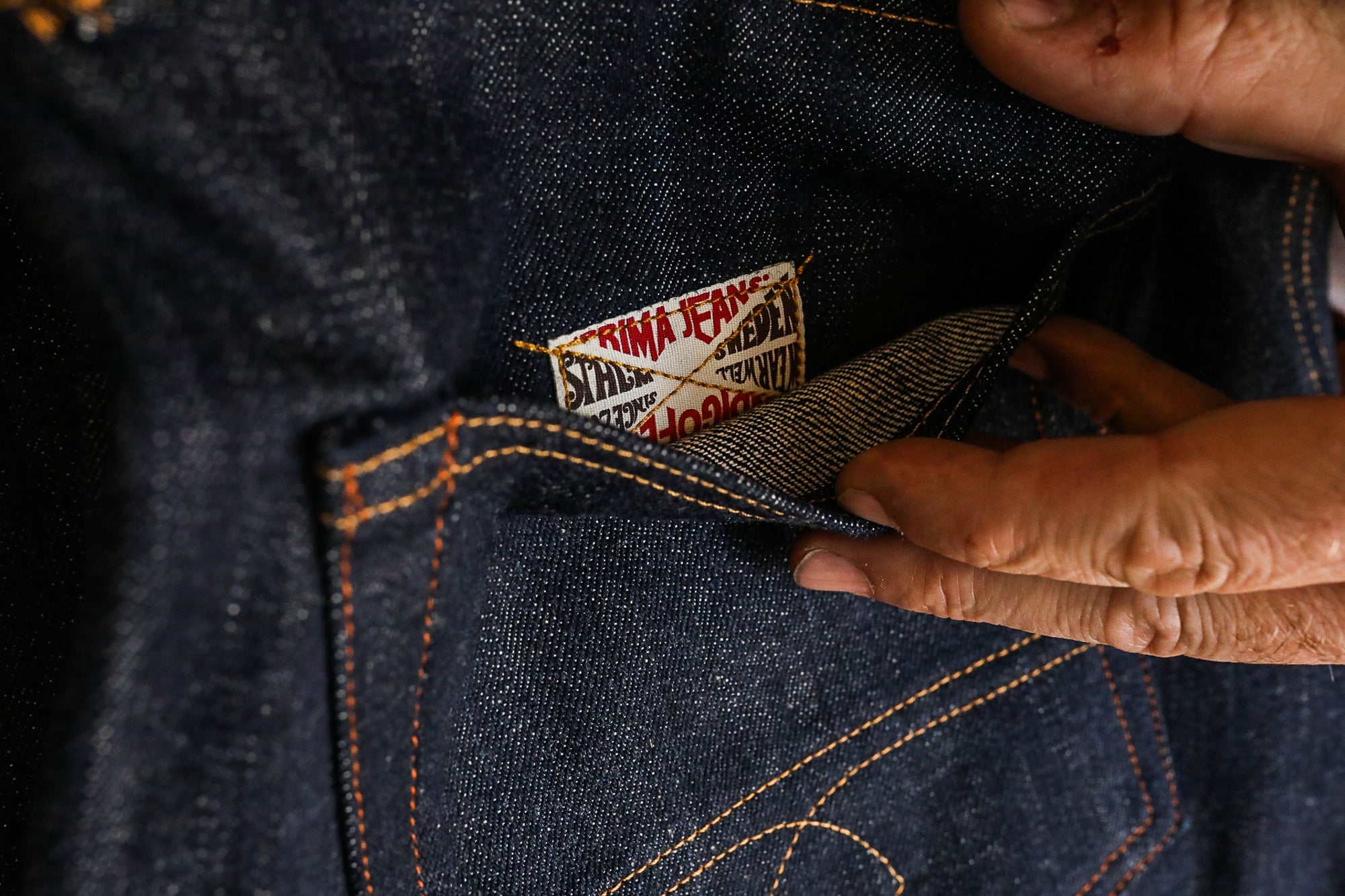Close-up of hands holding dark indigo denim pocket with visible Prima Jeans label and orange stitching