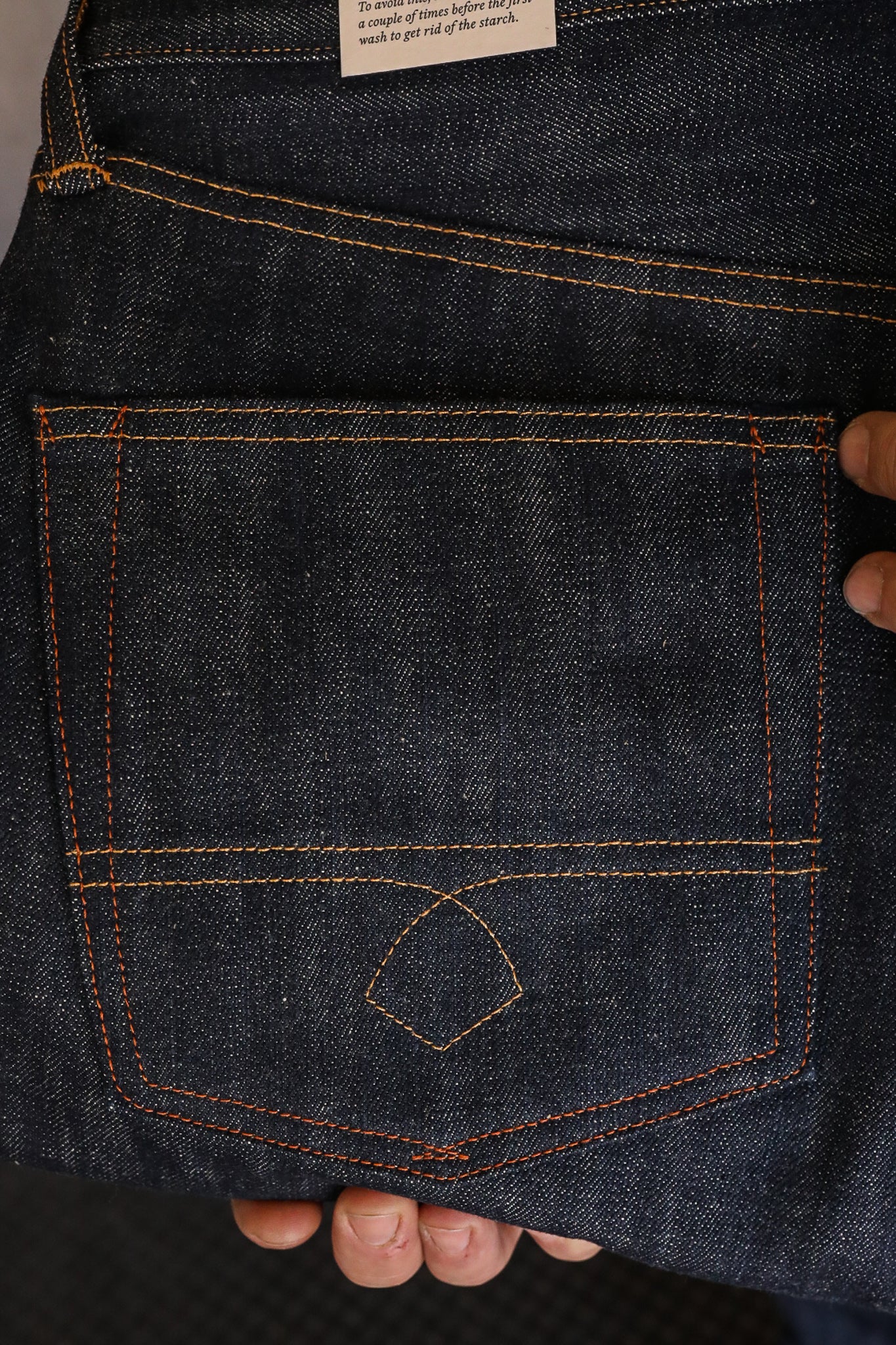 Close-up of dark denim jeans back pocket with orange stitching held by fingers, Iron Shop Provisions