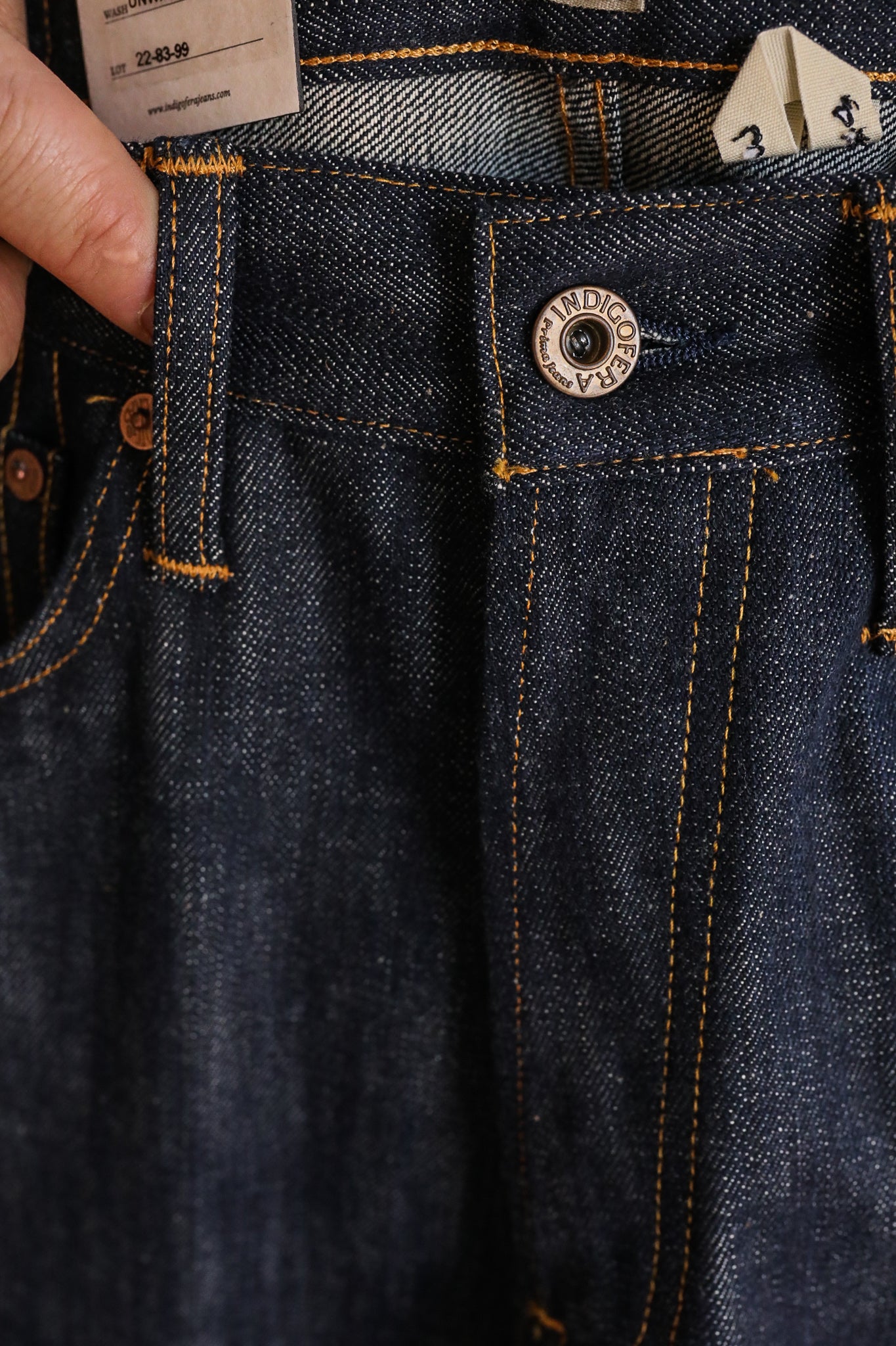 Close-up of dark indigo denim jeans with contrasting orange stitching and a metal button labeled 'Indigoeff'