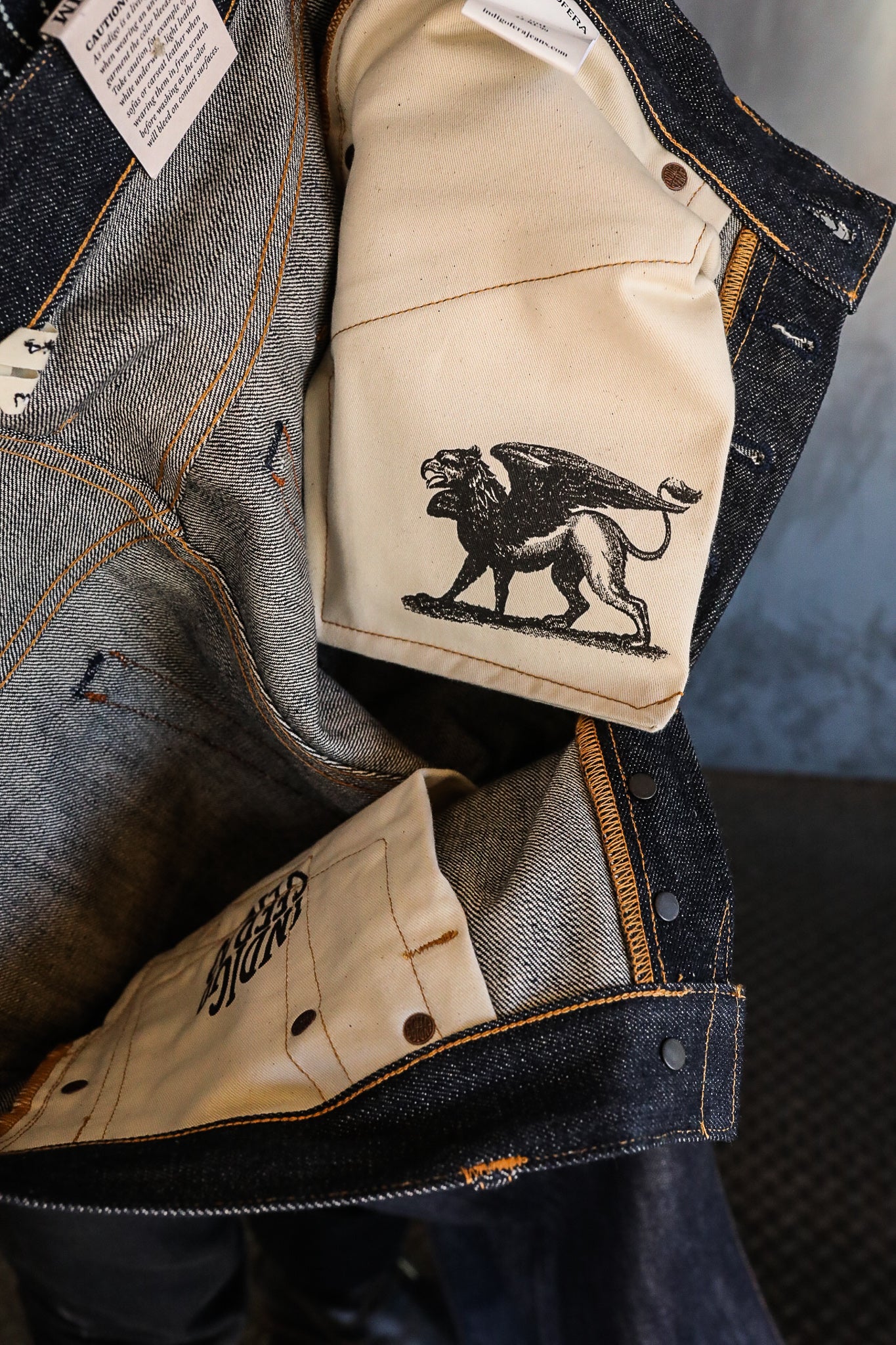 Interior of dark denim jeans showing beige pocket lining with winged griffin print and detailed stitching