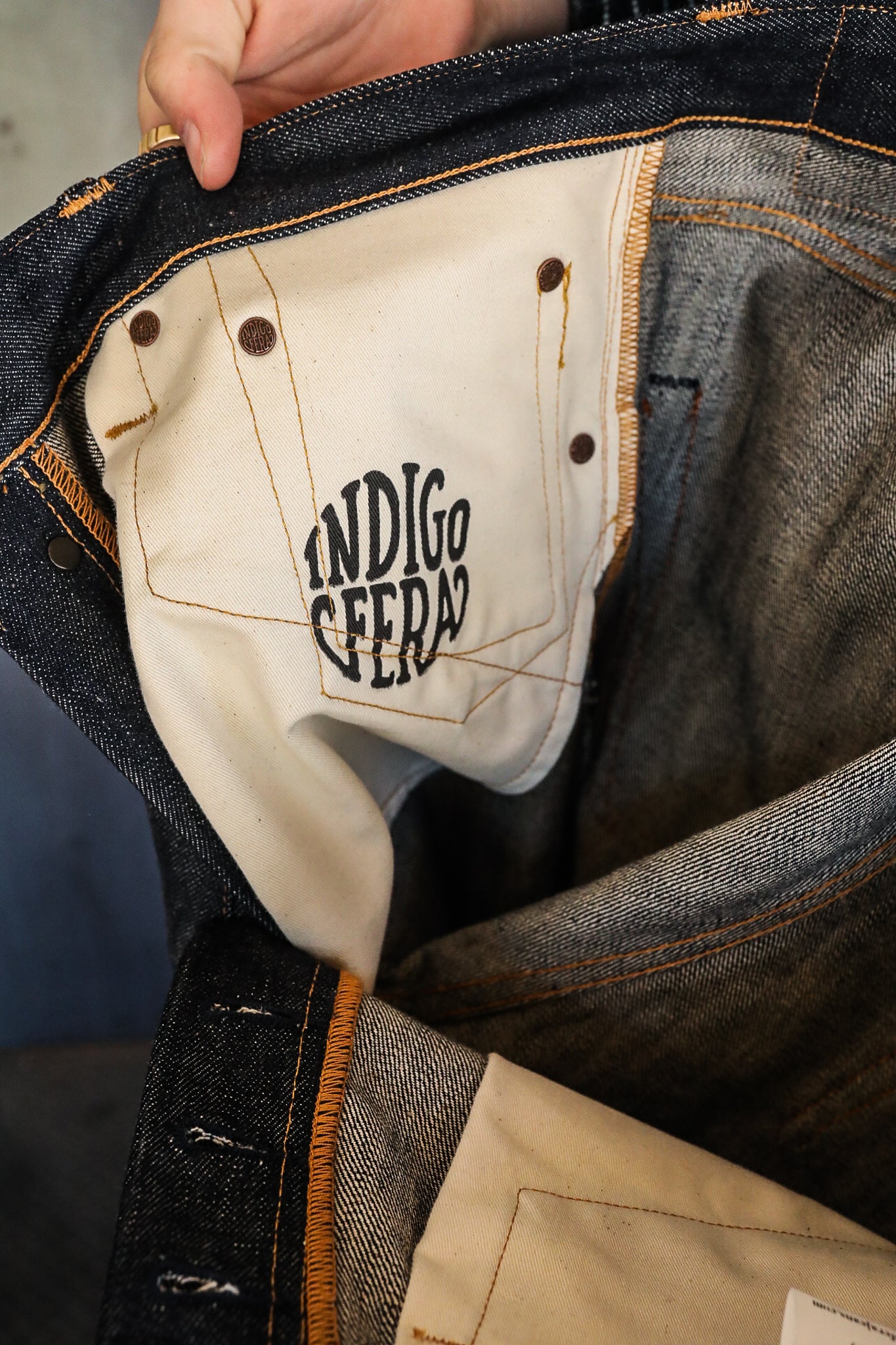 Hand holding open dark denim jeans showing white pocket lining with Indigofera logo and orange stitching