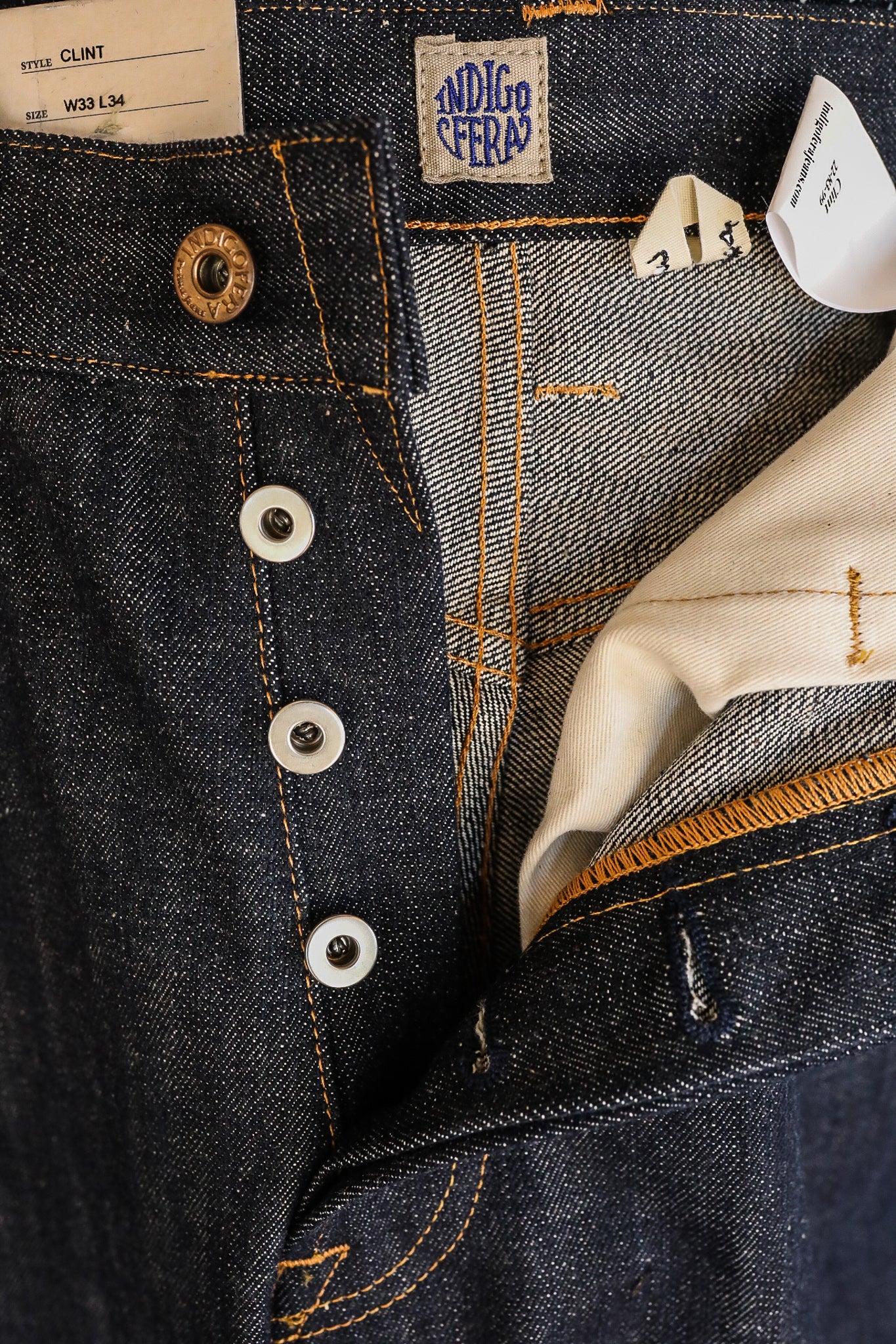 Close-up of Indigofera Clint dark blue denim jeans button fly with orange stitching and label