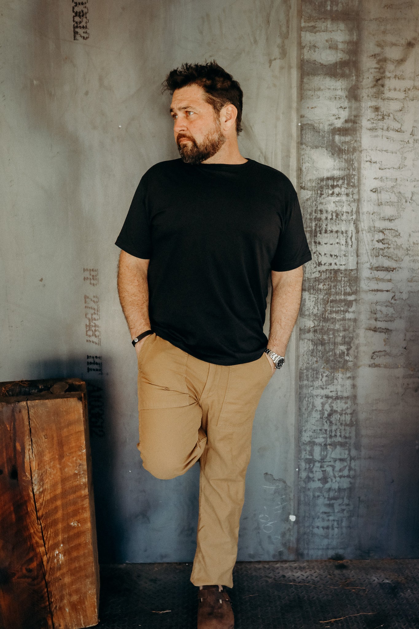 Man wearing black t-shirt and khaki pants leaning against industrial metal wall with wooden block