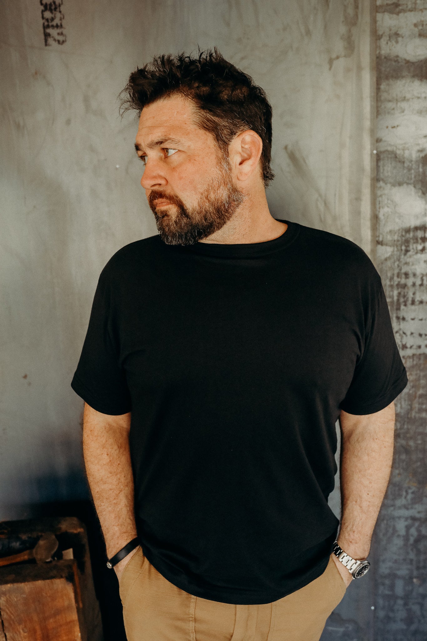 Bearded man wearing a black t-shirt and khaki pants standing against a textured industrial wall