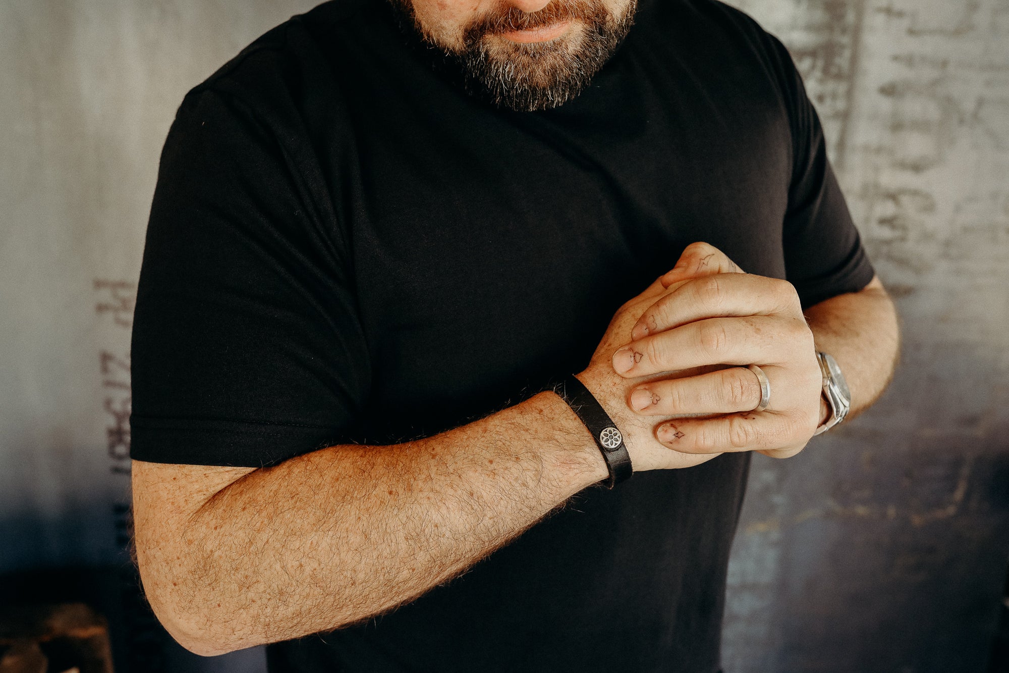 Man wearing black t-shirt and leather bracelet with metal emblem, showing hands with rings and tattoos