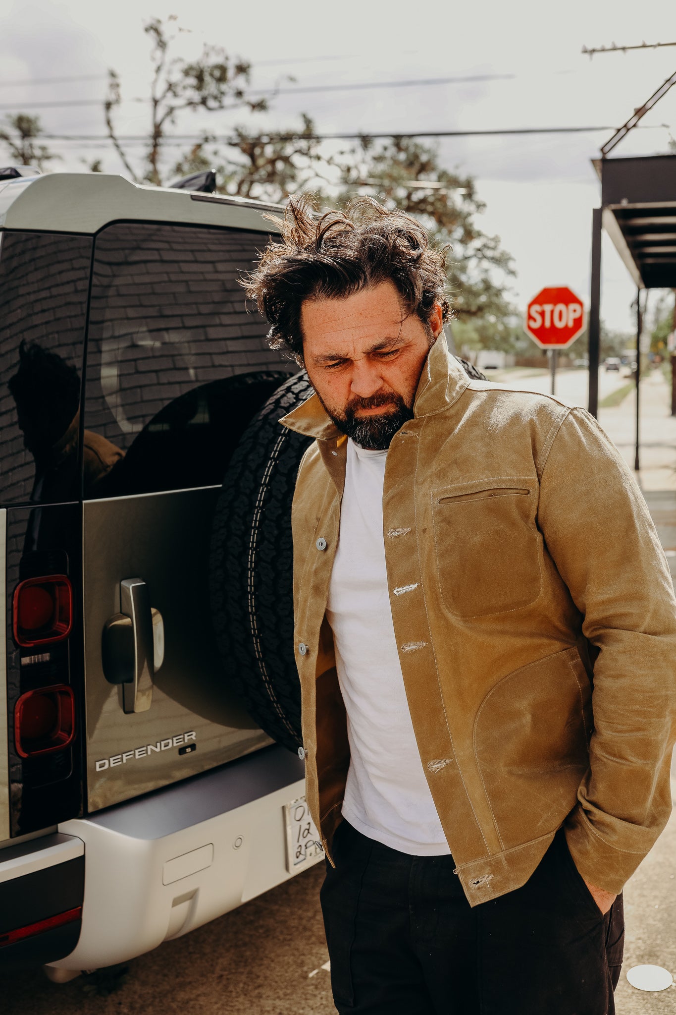Man wearing brown rugged jacket and white t-shirt leaning against a Land Rover Defender SUV on street