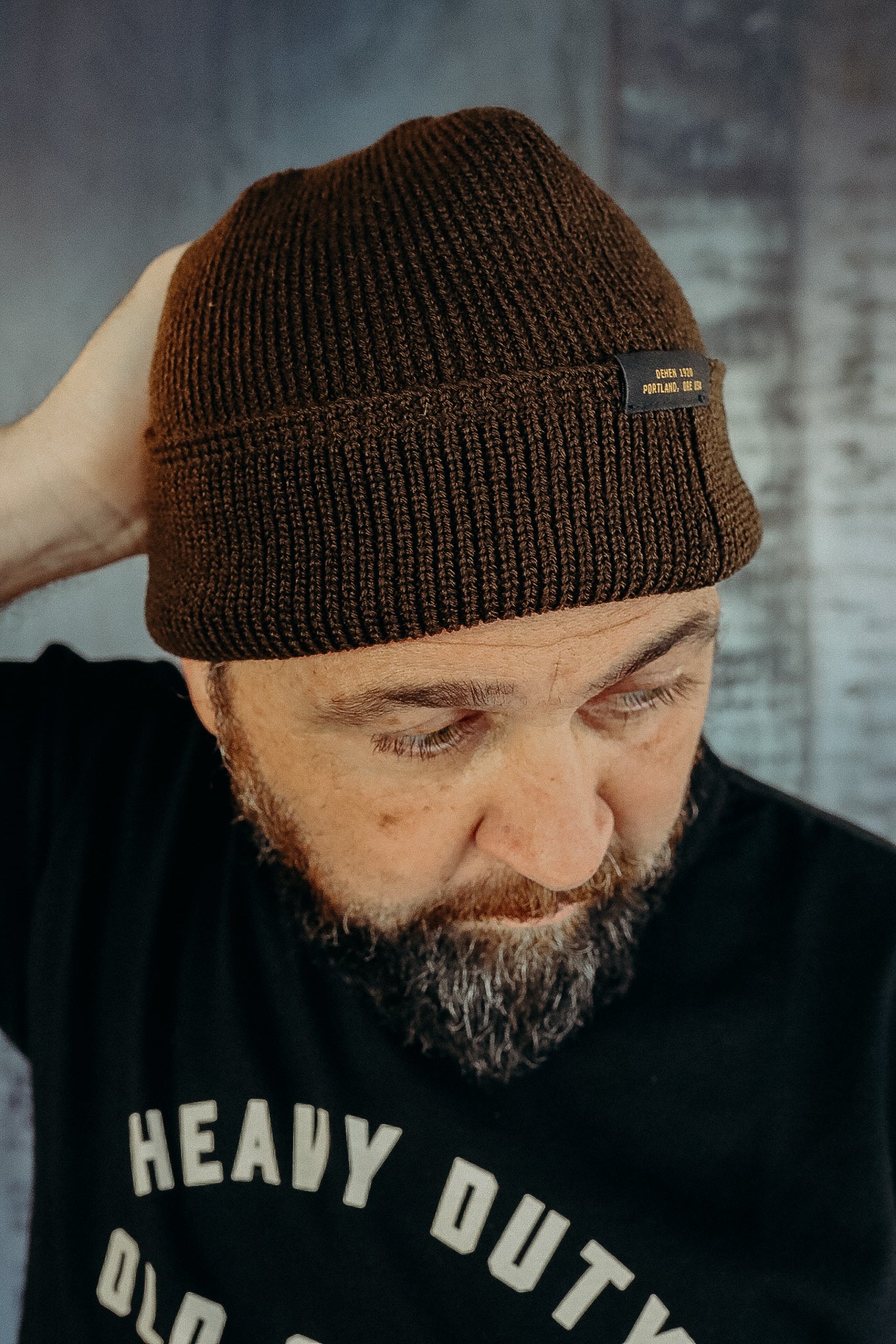 Man wearing brown knitted beanie with label, black shirt with heavy duty print, textured background