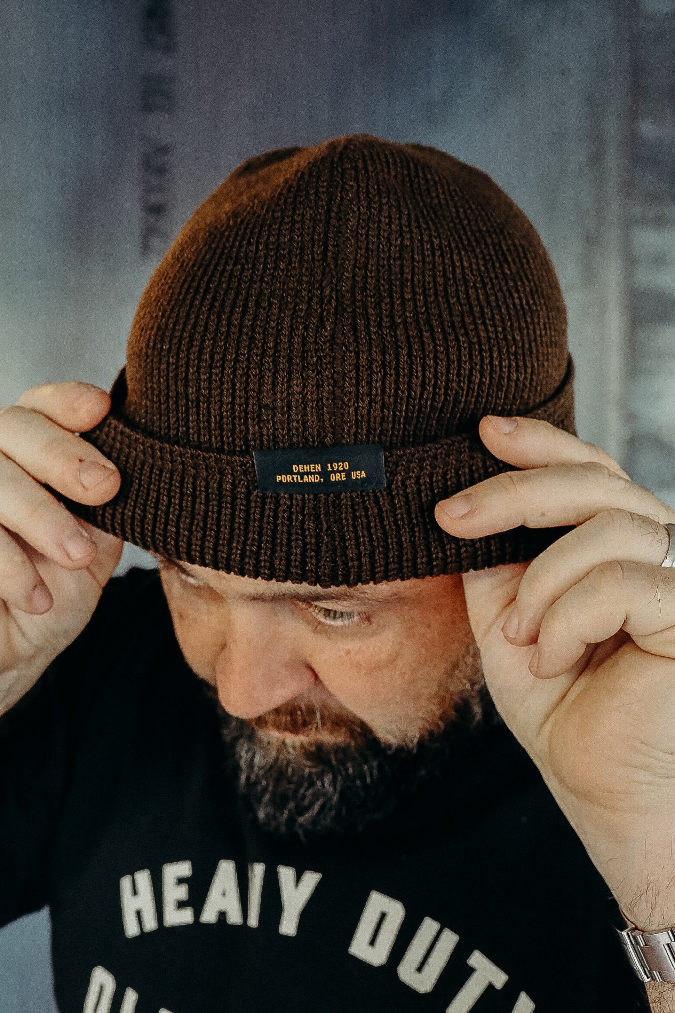 Man wearing brown knit beanie hat with Dehen 1920 Portland, Ore USA label, adjusting brim
