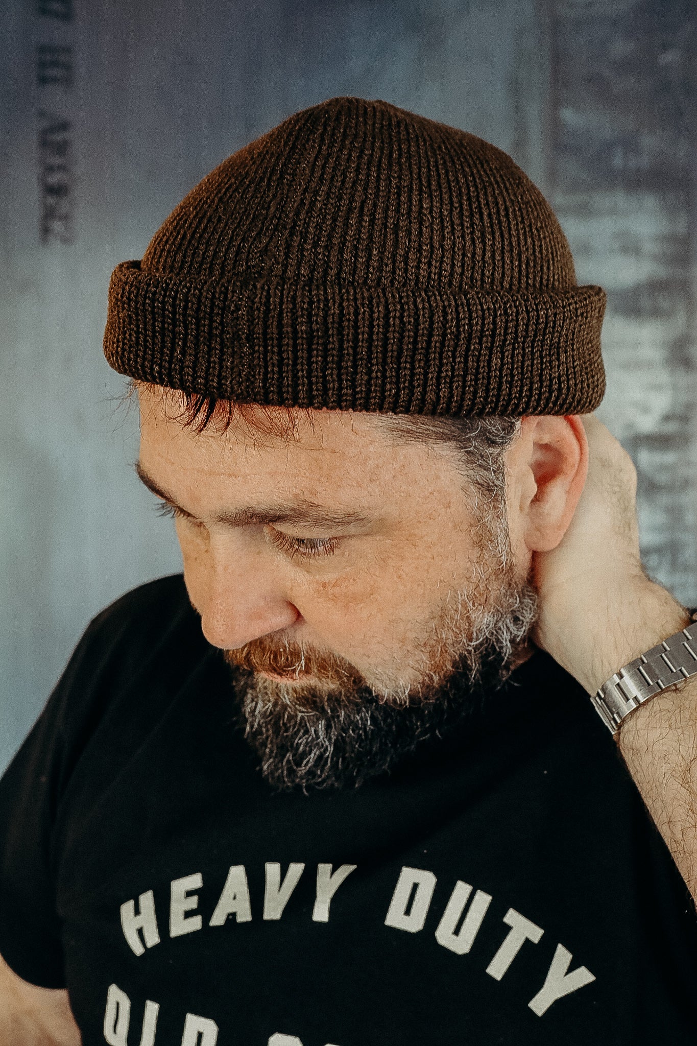 Man wearing brown knit beanie and black heavy duty t-shirt, looking down with hand behind neck
