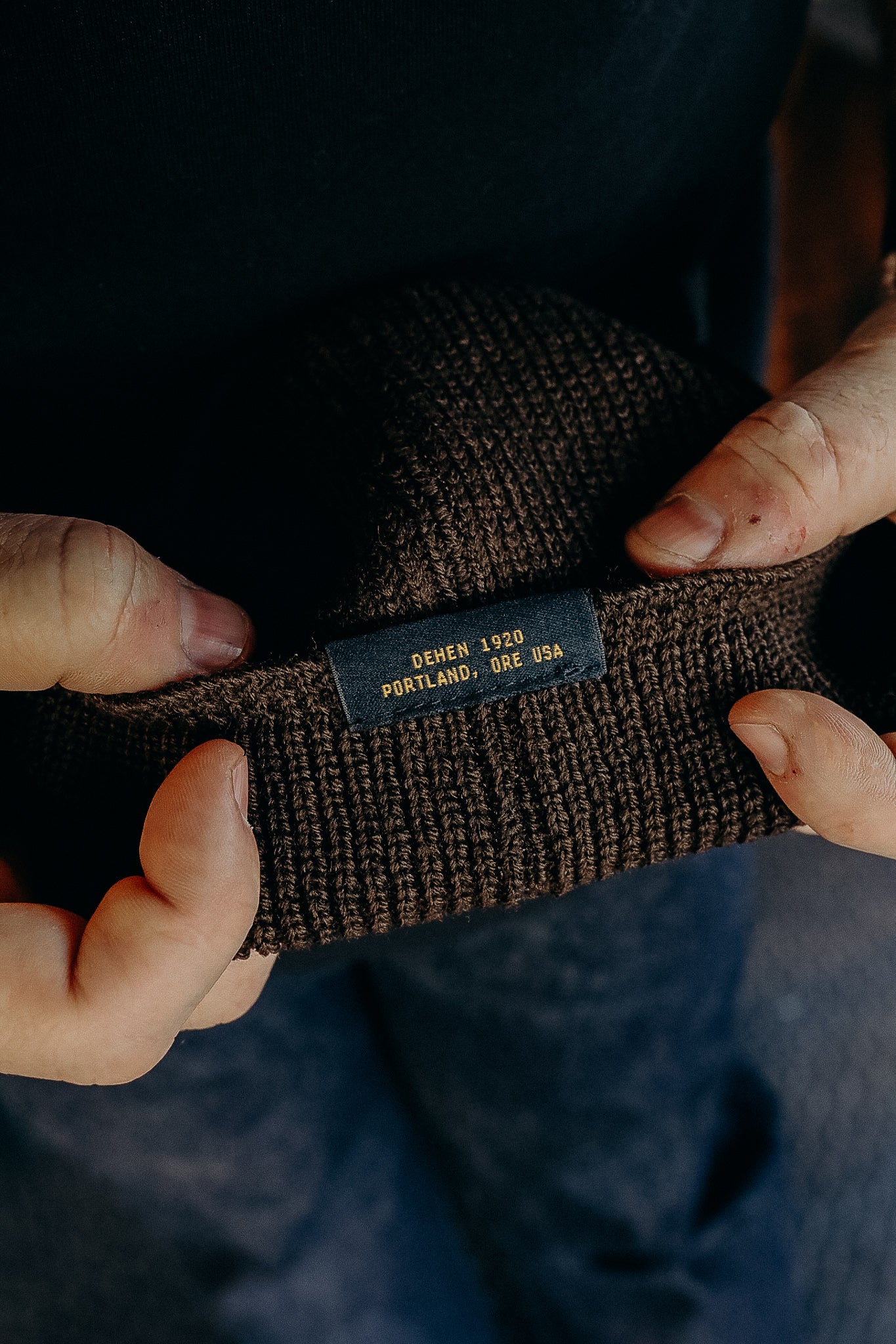Hands holding a brown knitted beanie with a label reading Dehen 1920 Portland, Ore USA, warm winter hat