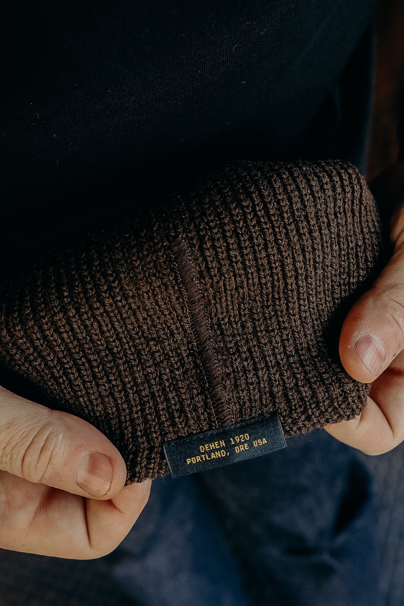 Close-up of hands holding a brown knitted beanie with Dehen 1920 Portland, Ore USA label