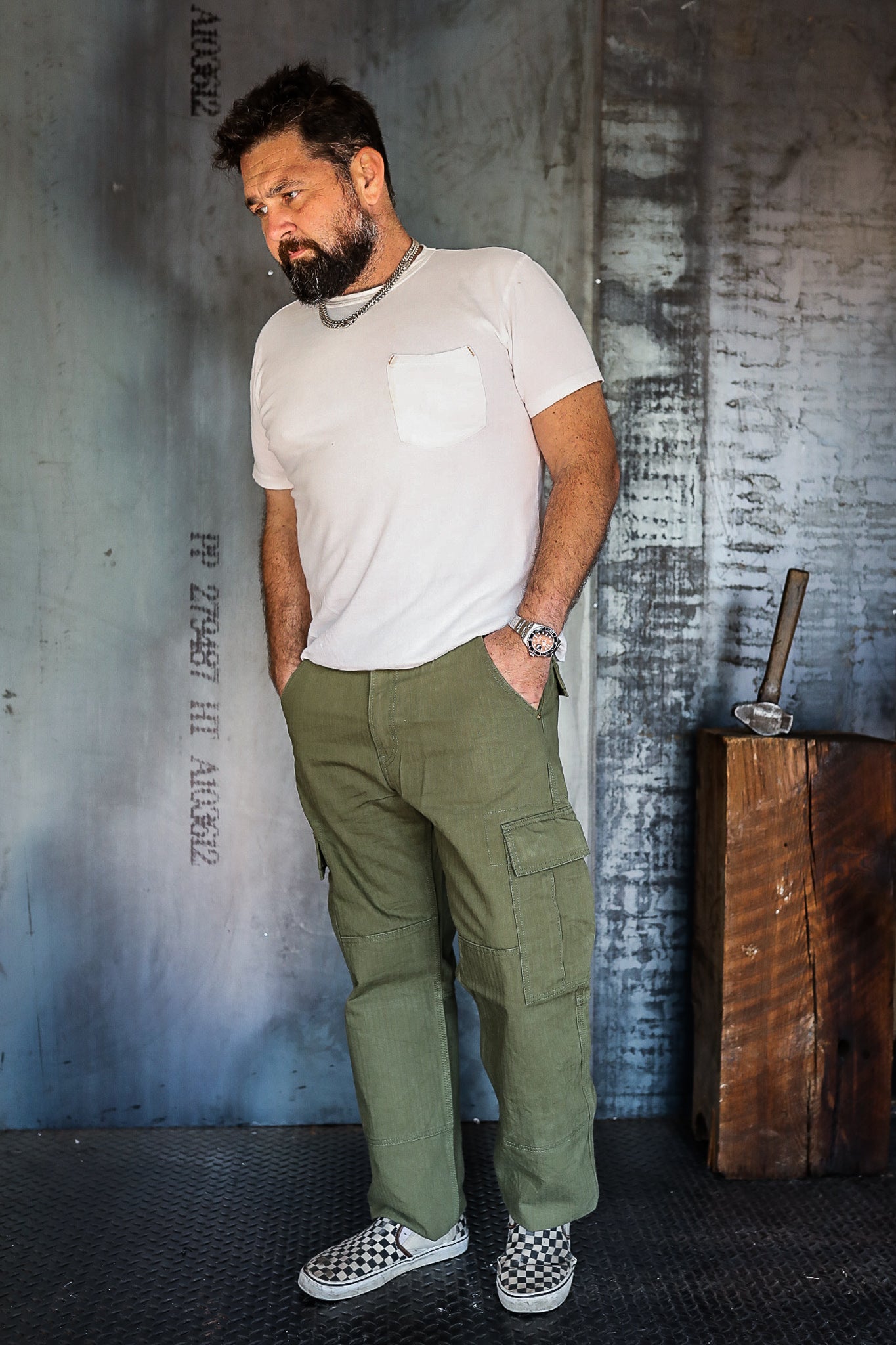 Bearded man in white pocket t-shirt, olive cargo pants, and checkered slip-on shoes standing in industrial setting with axe on wooden block