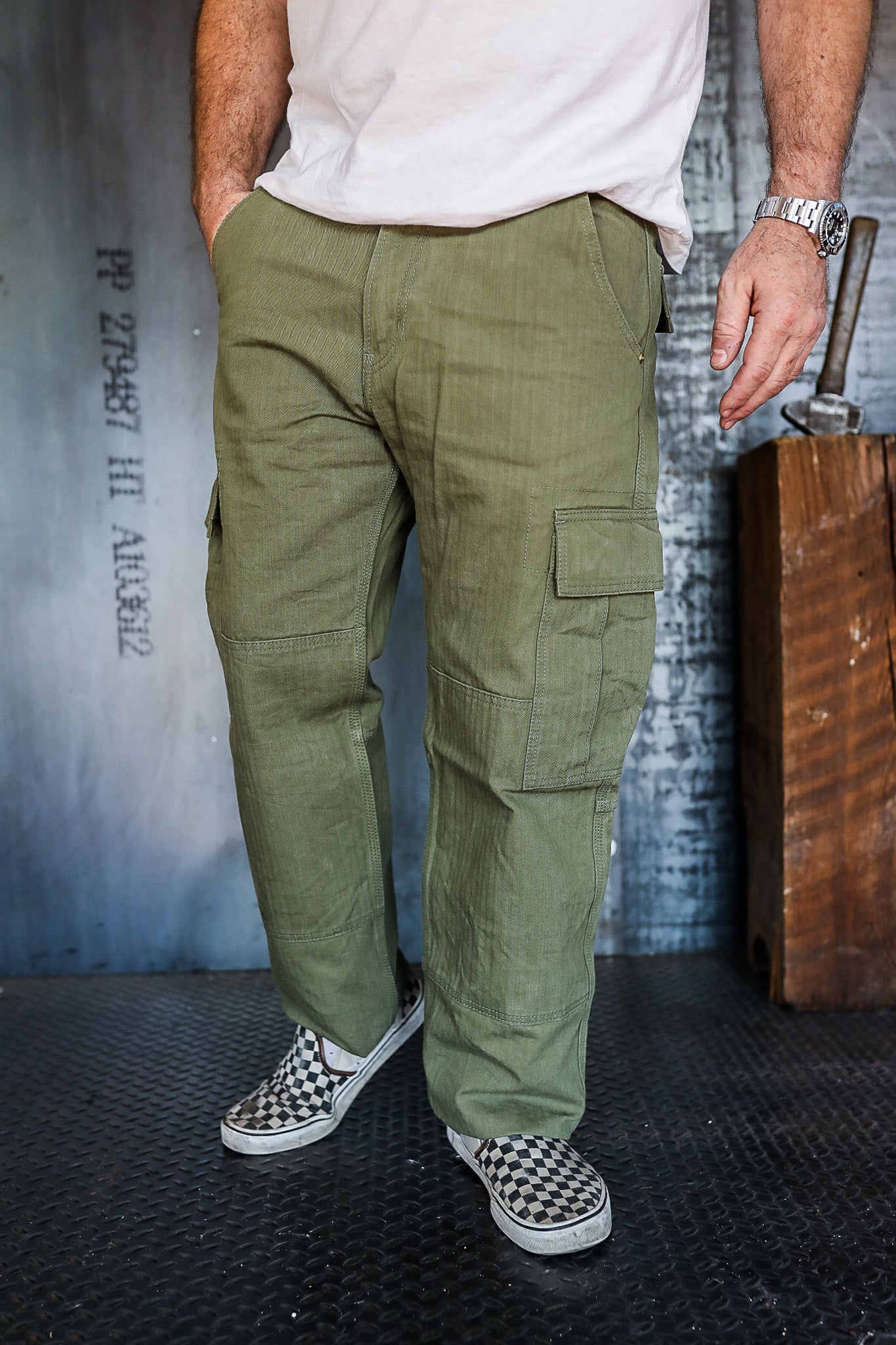 Man wearing olive green cargo pants and checkered slip-on shoes standing on textured black floor
