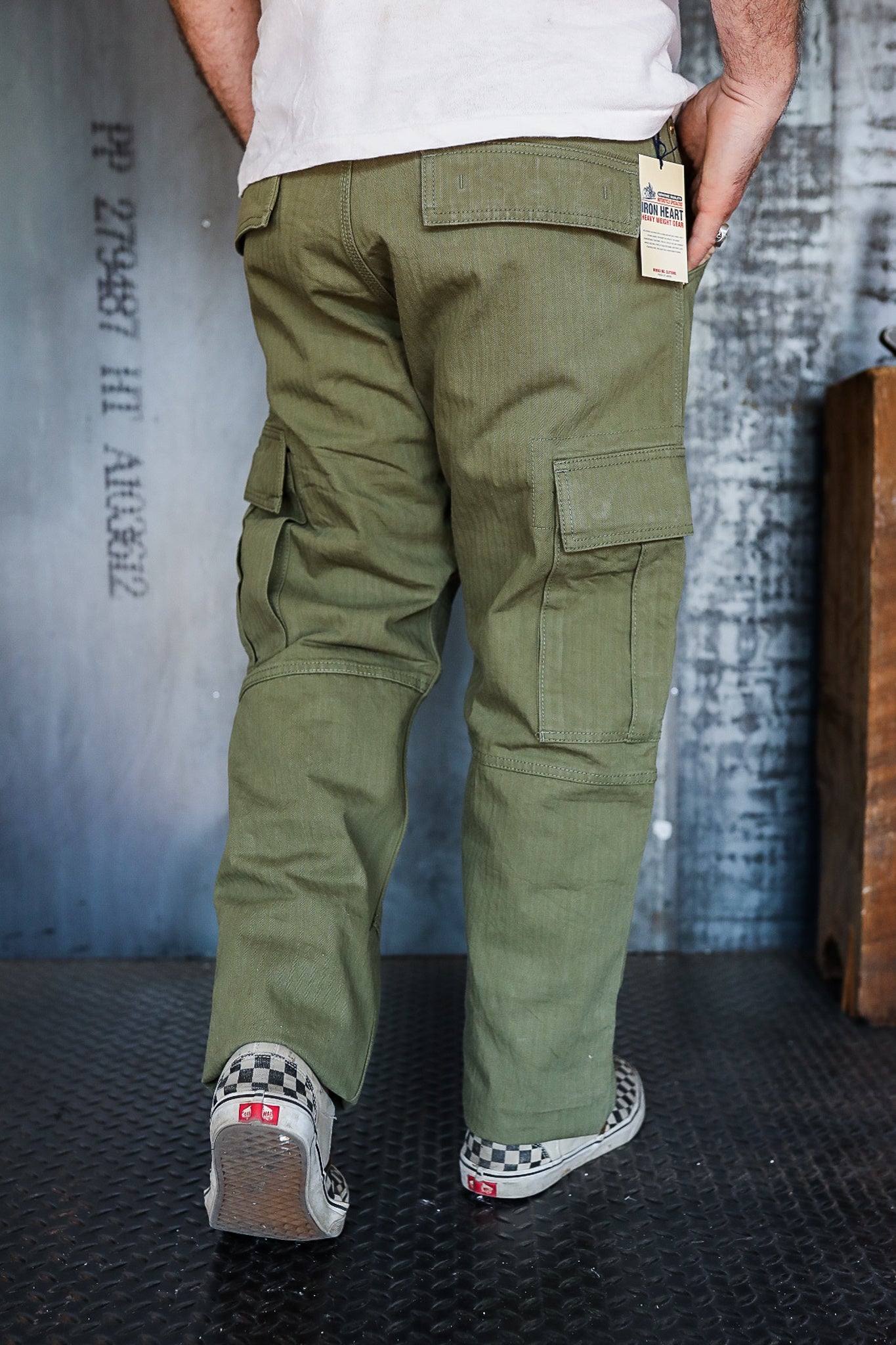 Rear view of a person wearing olive green cargo pants with rear and side flap pockets and checkerboard slip-on sneakers