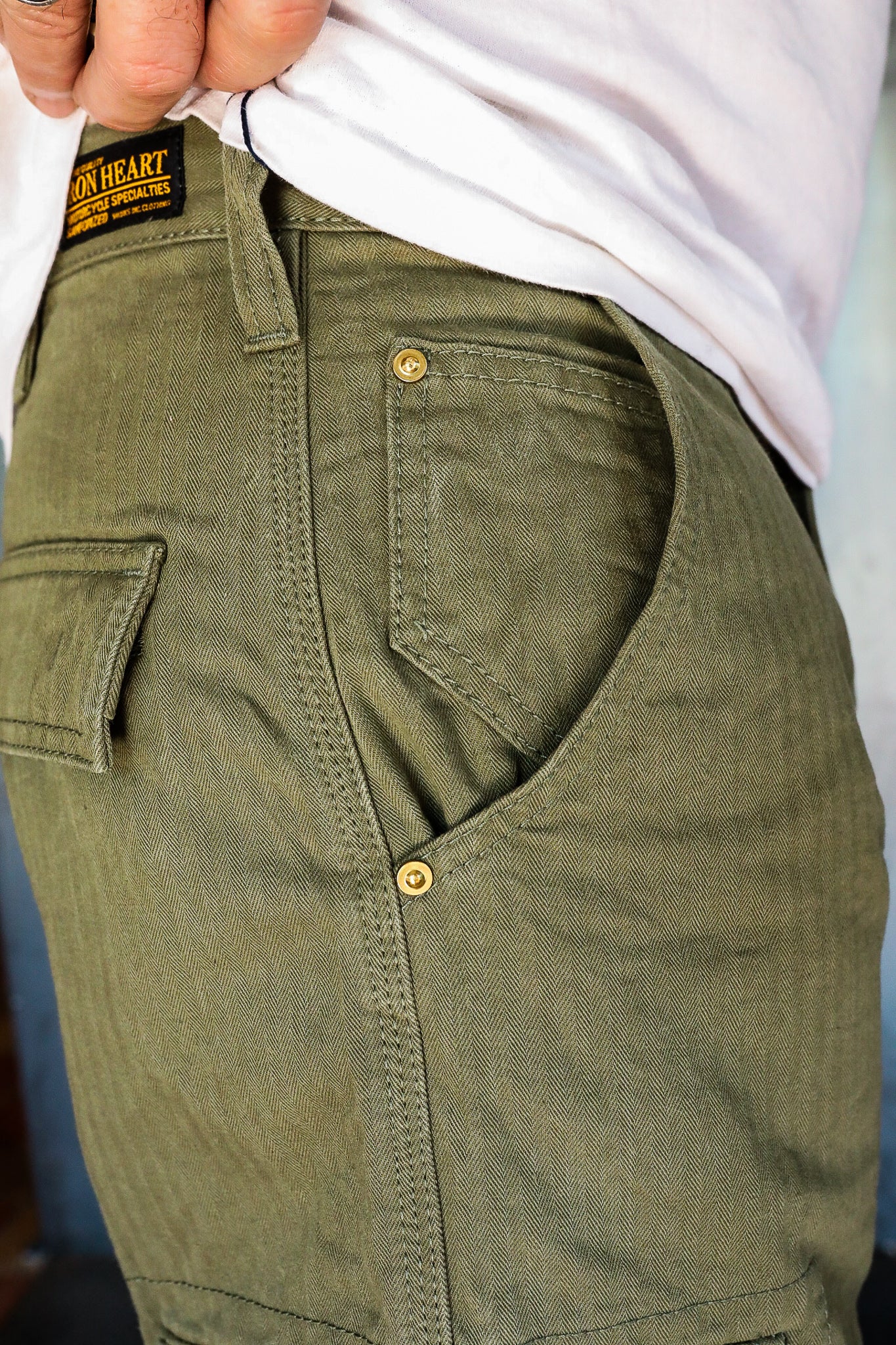 Close-up of olive green herringbone work pants with brass rivets and Iron Heart brand patch