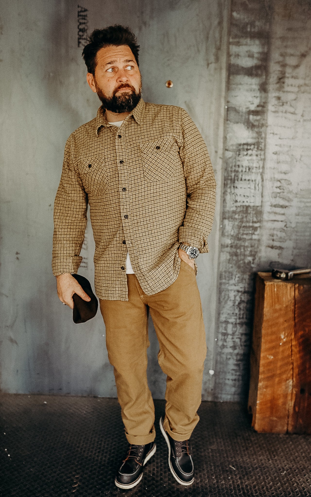 Man wearing plaid button-up shirt, tan pants, and boots, standing in industrial setting at Iron Shop Provisions