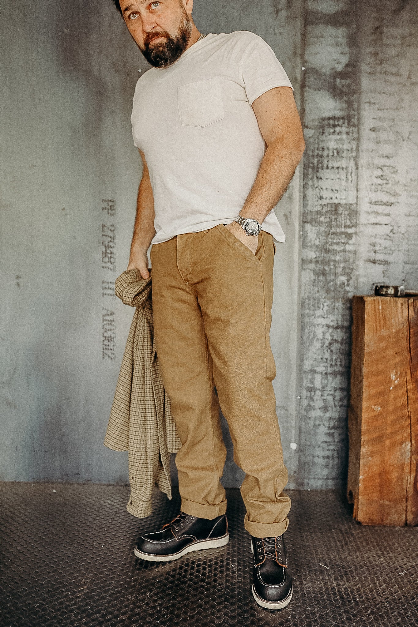 Man wearing beige pants and dark work boots holding a plaid jacket in an industrial setting