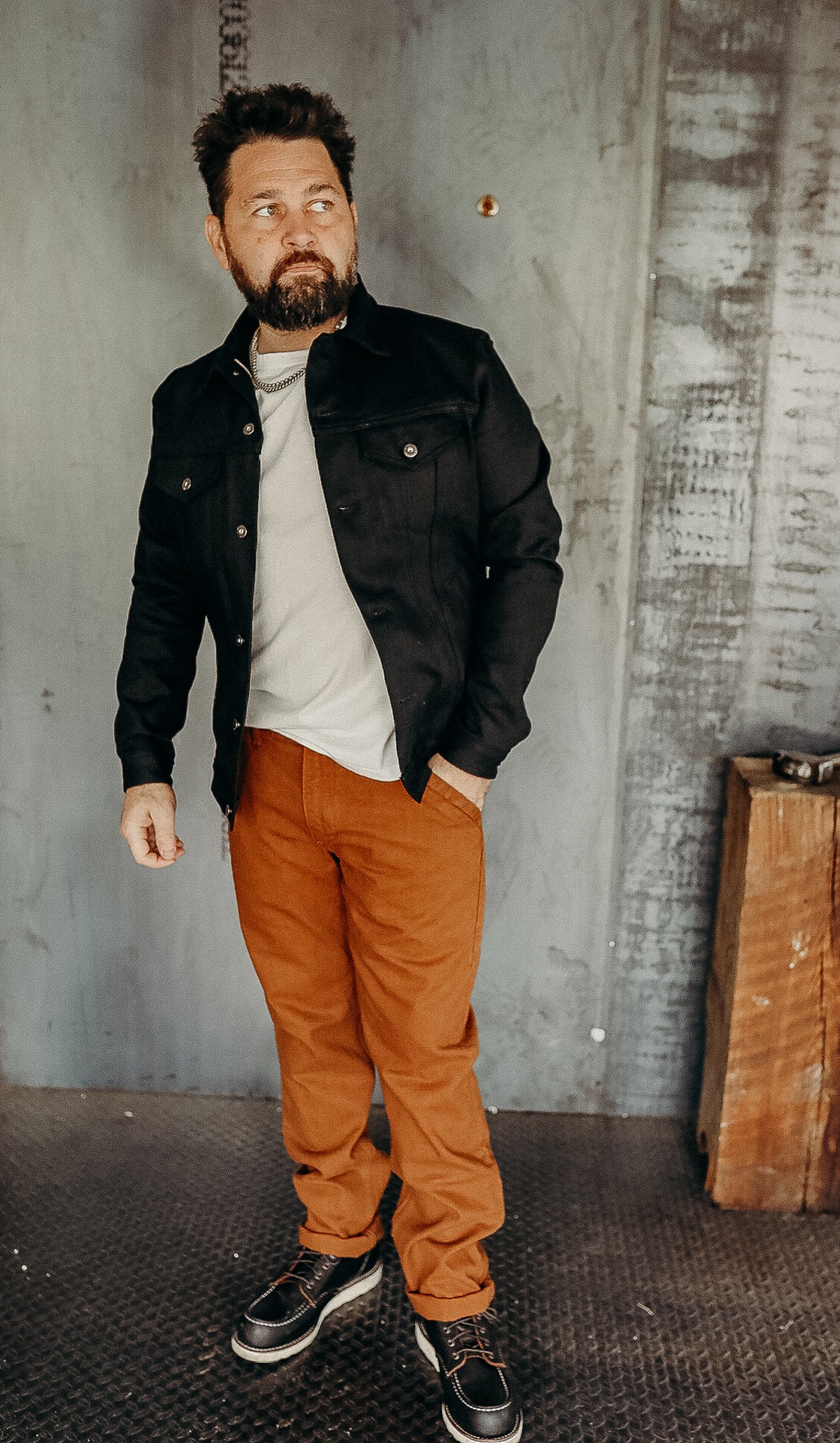 Man wearing black jacket, white shirt, brown pants, and black work boots in industrial setting