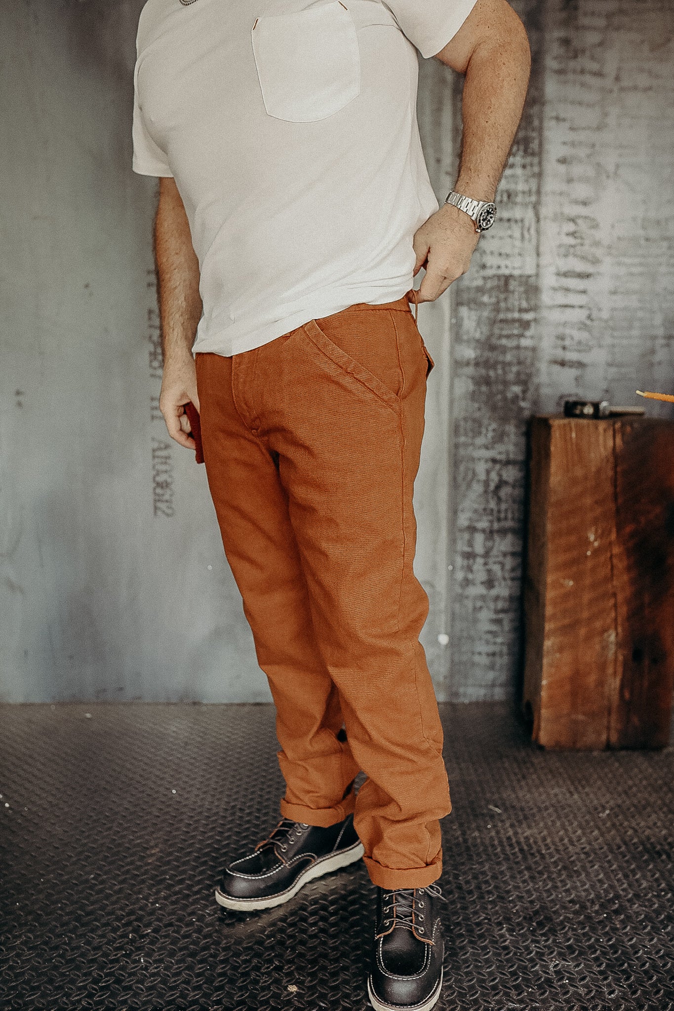 Man wearing rust-colored work pants, white t-shirt, and black leather boots standing on metal floor