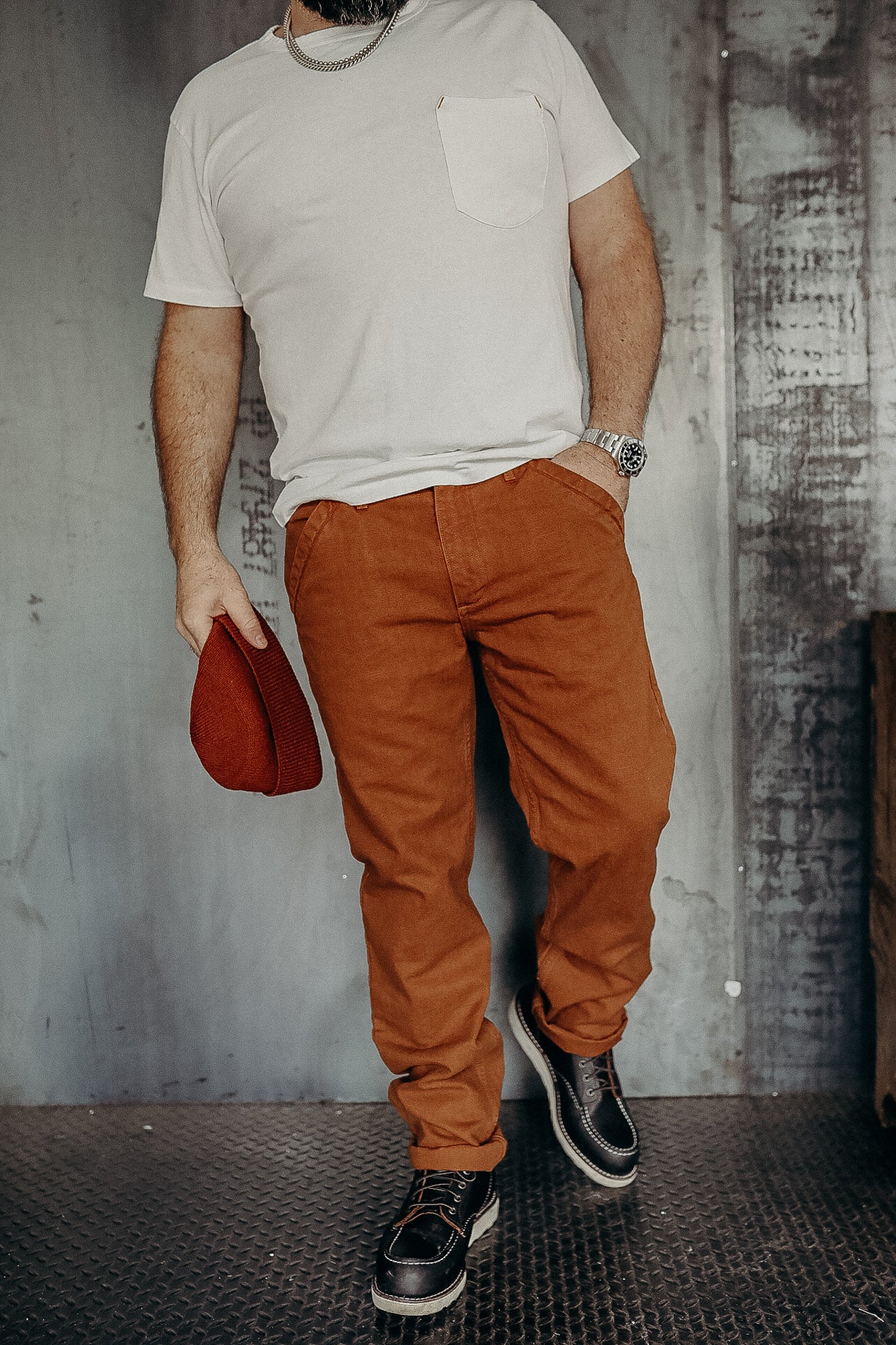 Man wearing white pocket t-shirt, rust-colored pants, black leather boots, holding red knit hat in industrial setting