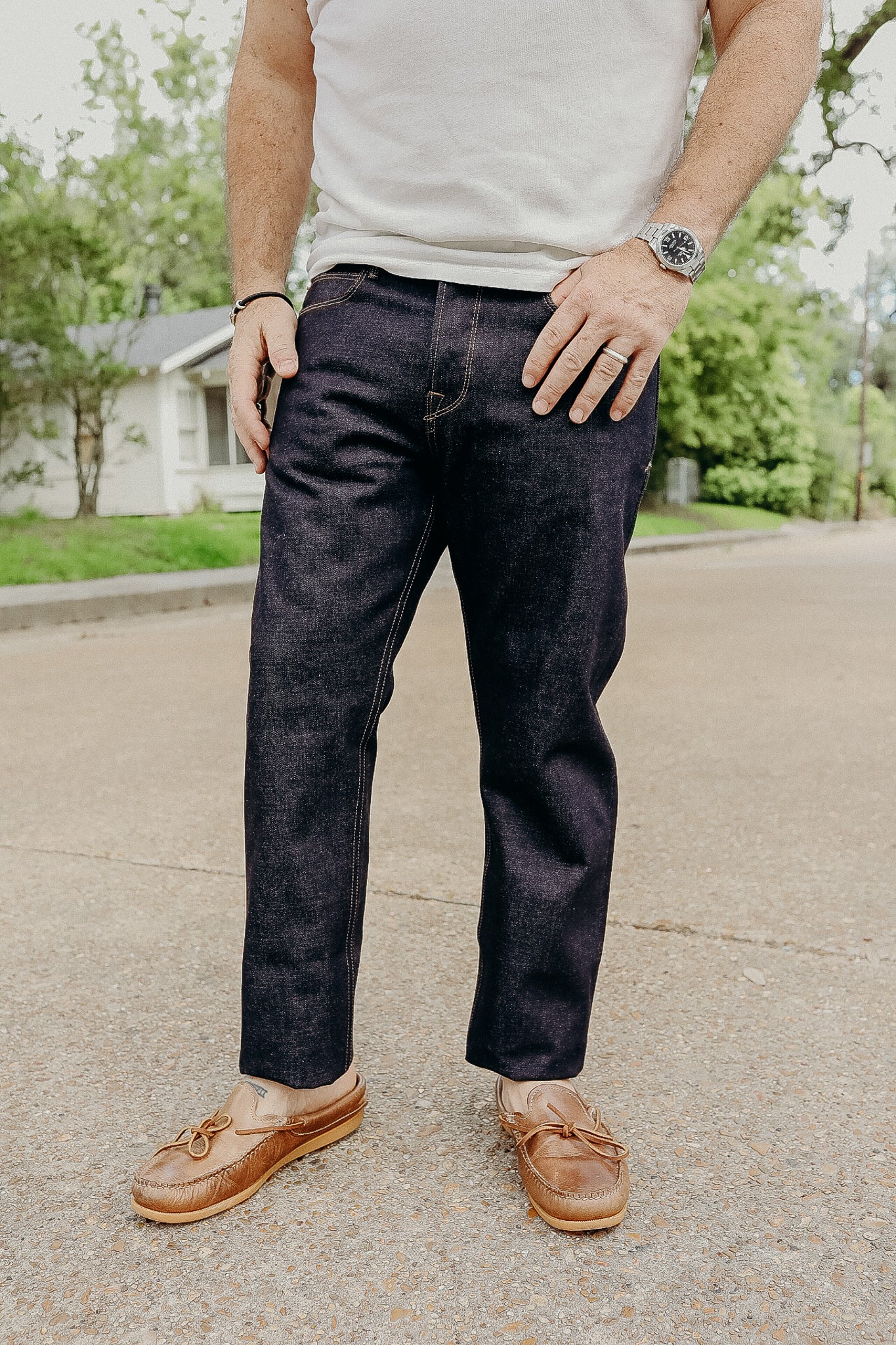 Man wearing dark raw denim jeans, white t-shirt, brown leather moccasins, standing outdoors on street