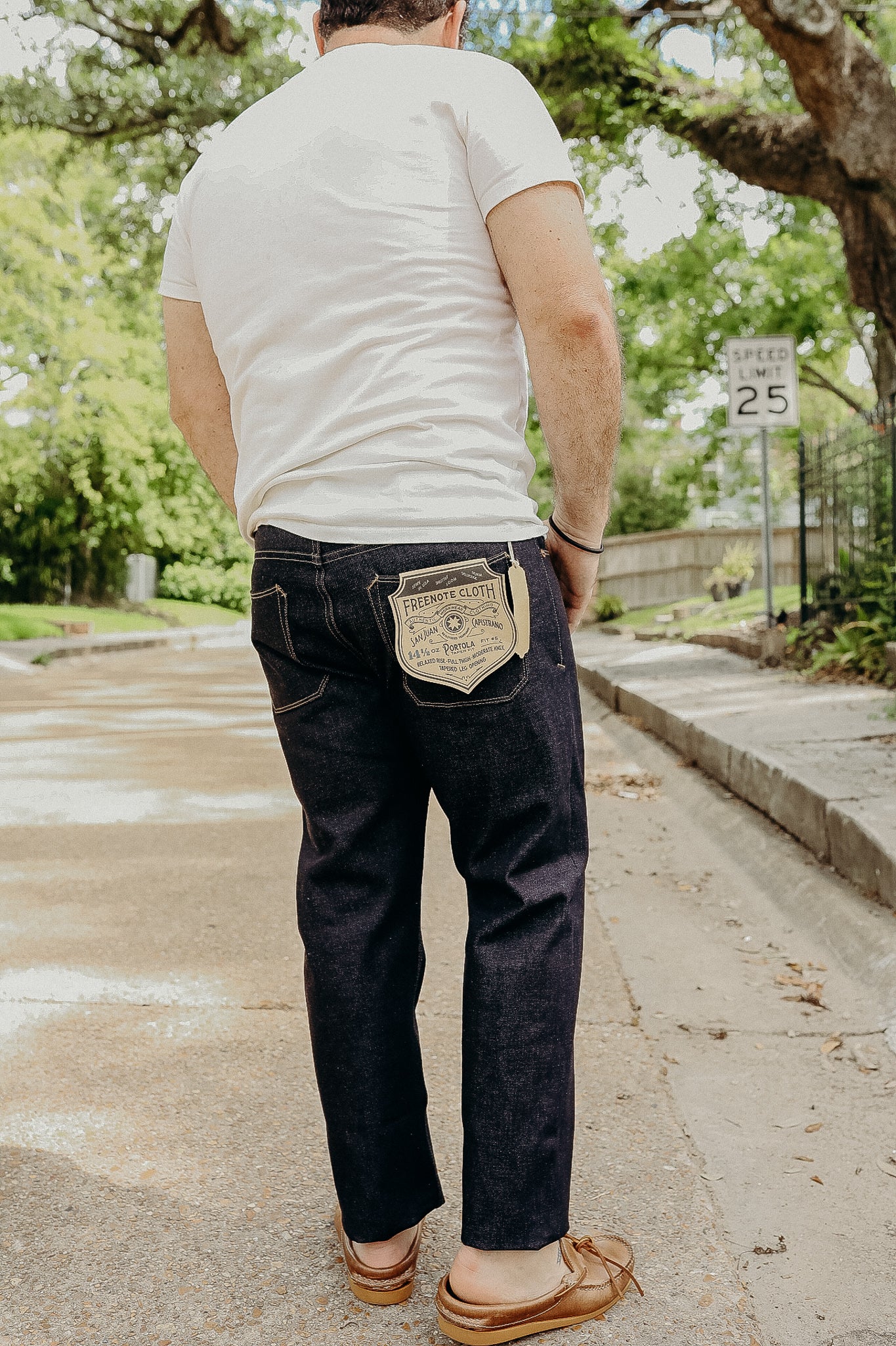 Man wearing dark denim jeans with Freenote Cloth patch and white t-shirt walking on a tree-lined street