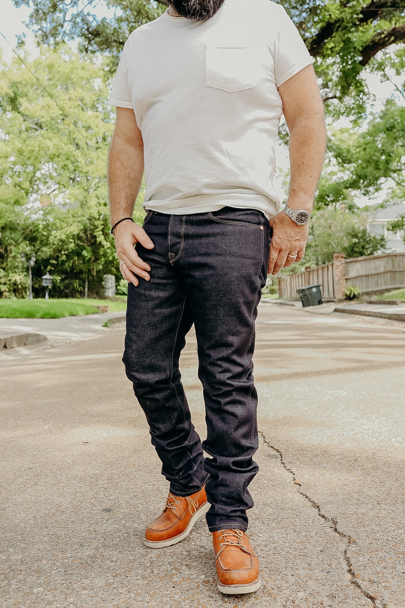 Man standing outdoors wearing dark denim jeans, white t-shirt, brown leather boots, and watch