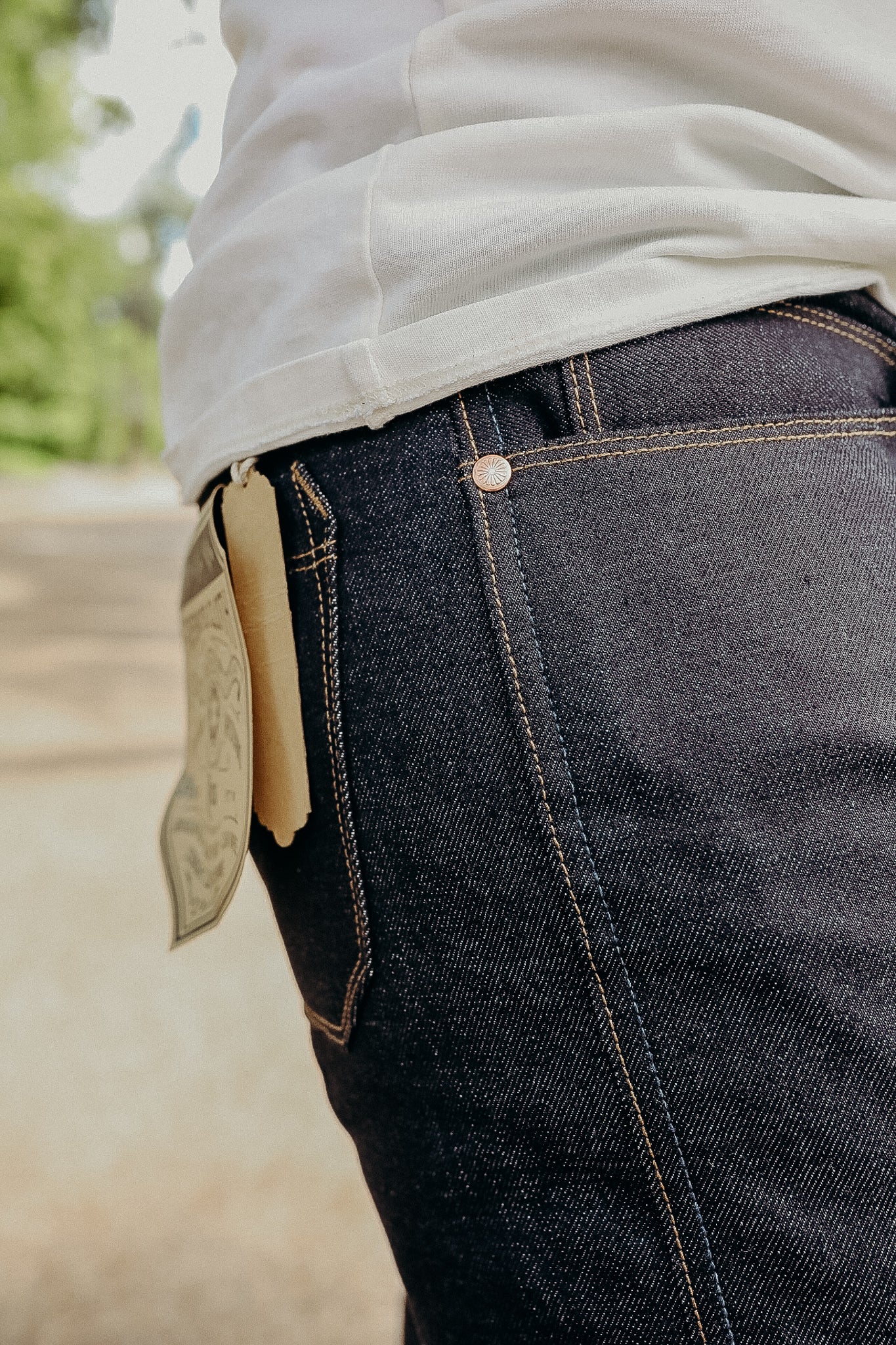 Close-up of dark denim jeans with tan stitching worn with a white shirt outdoors, showing a paper tag