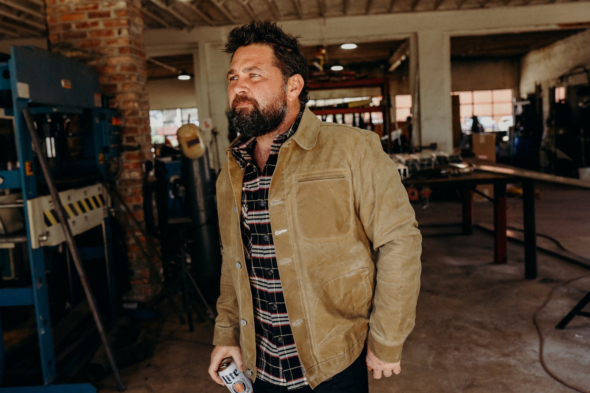 Bearded man in a tan work jacket and plaid shirt standing in an industrial workshop holding a can of beer