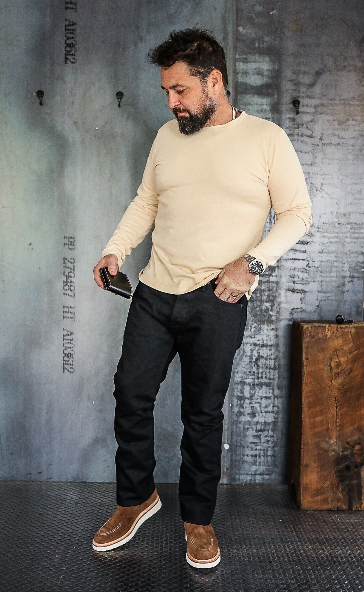 Bearded man in beige long-sleeve shirt, black pants, and brown shoes standing indoors with industrial wall background