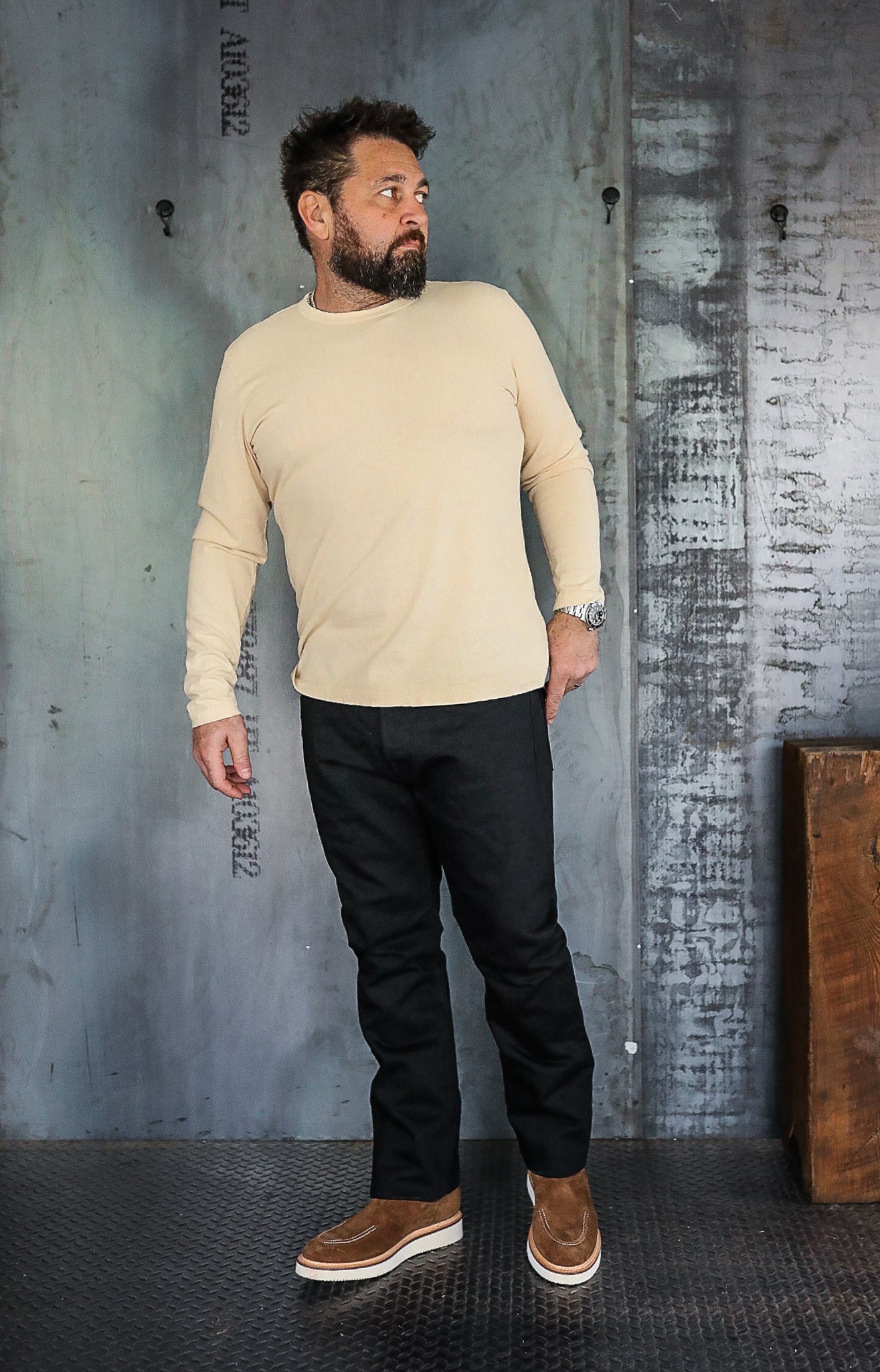 Man wearing beige long-sleeve shirt, black pants, and brown suede shoes in industrial setting
