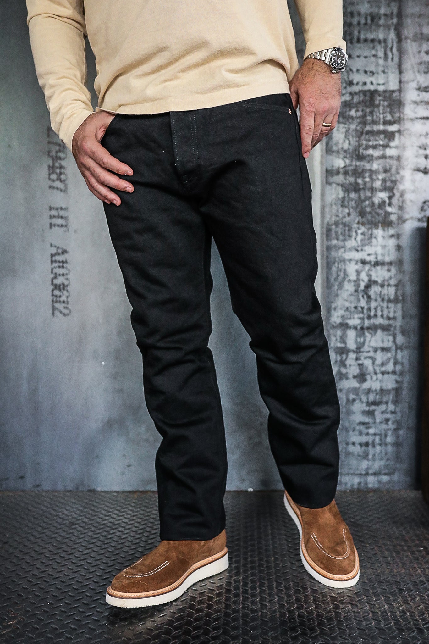 Man wearing black work pants and tan suede boots standing on textured metal floor in industrial setting