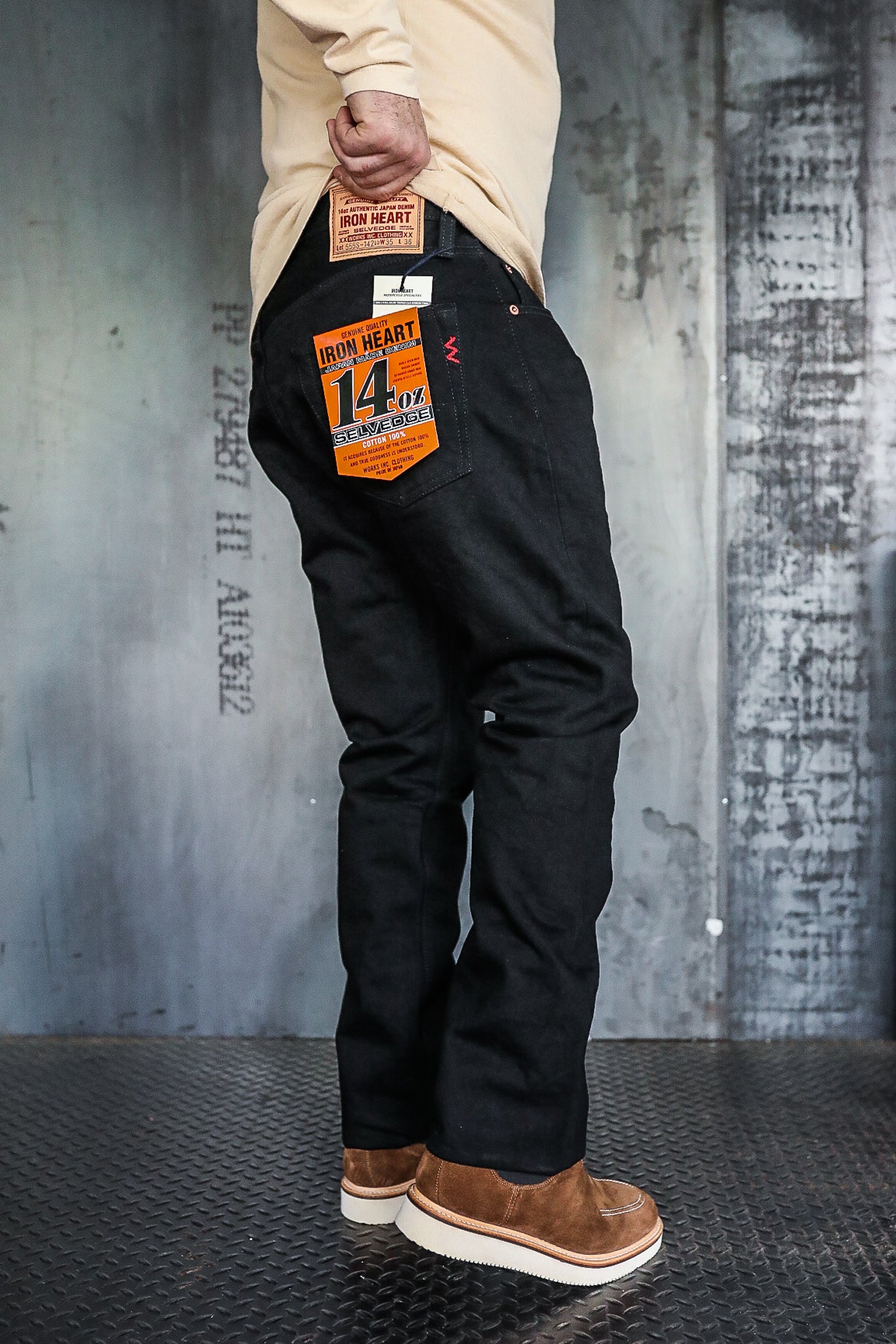 Man wearing black Iron Heart 14 oz selvedge denim jeans with tan suede shoes against industrial backdrop