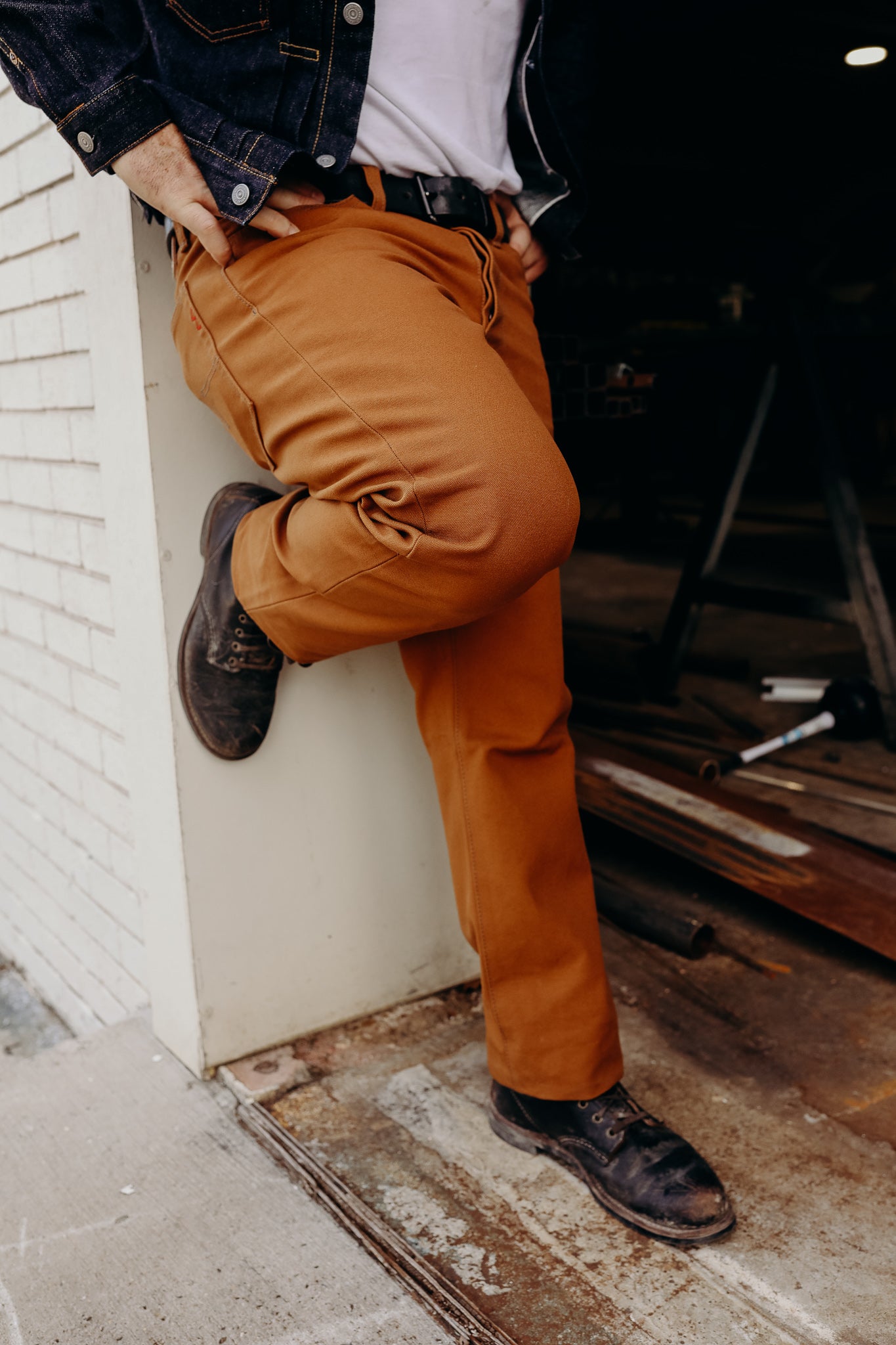 Person wearing durable brown work pants, dark leather boots, and denim jacket, leaning against white brick wall