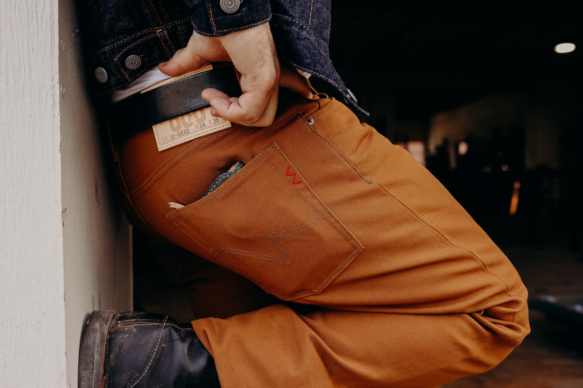 Close-up of person wearing brown work pants with belt and denim jacket, kneeling on rough floor indoors
