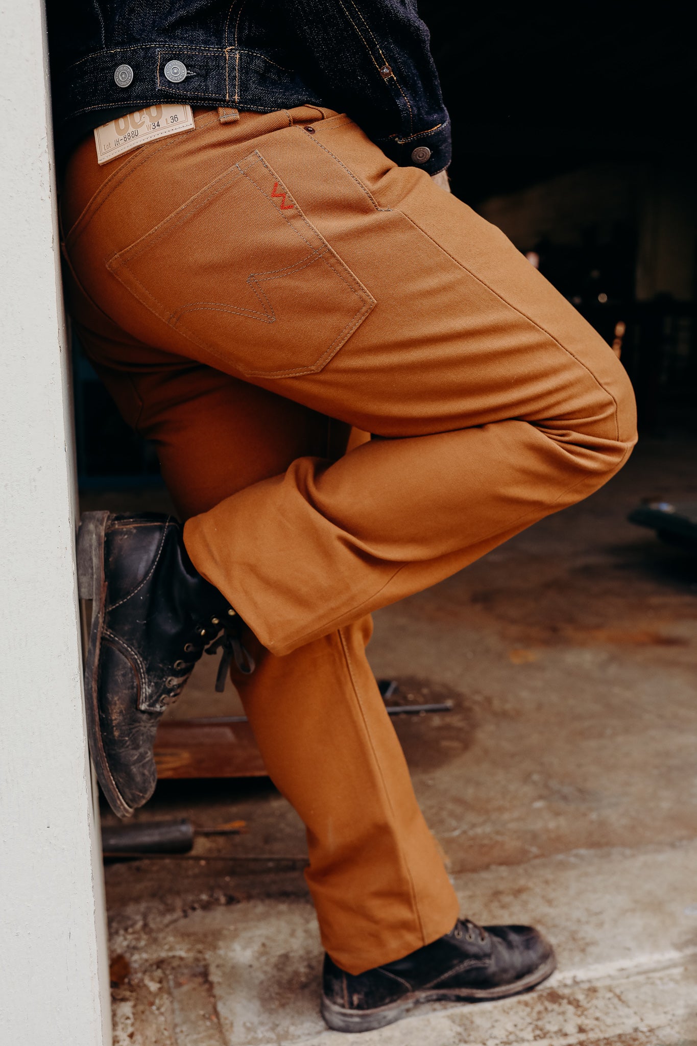 Person wearing brown work pants and worn black leather boots leaning against a wall in a workshop