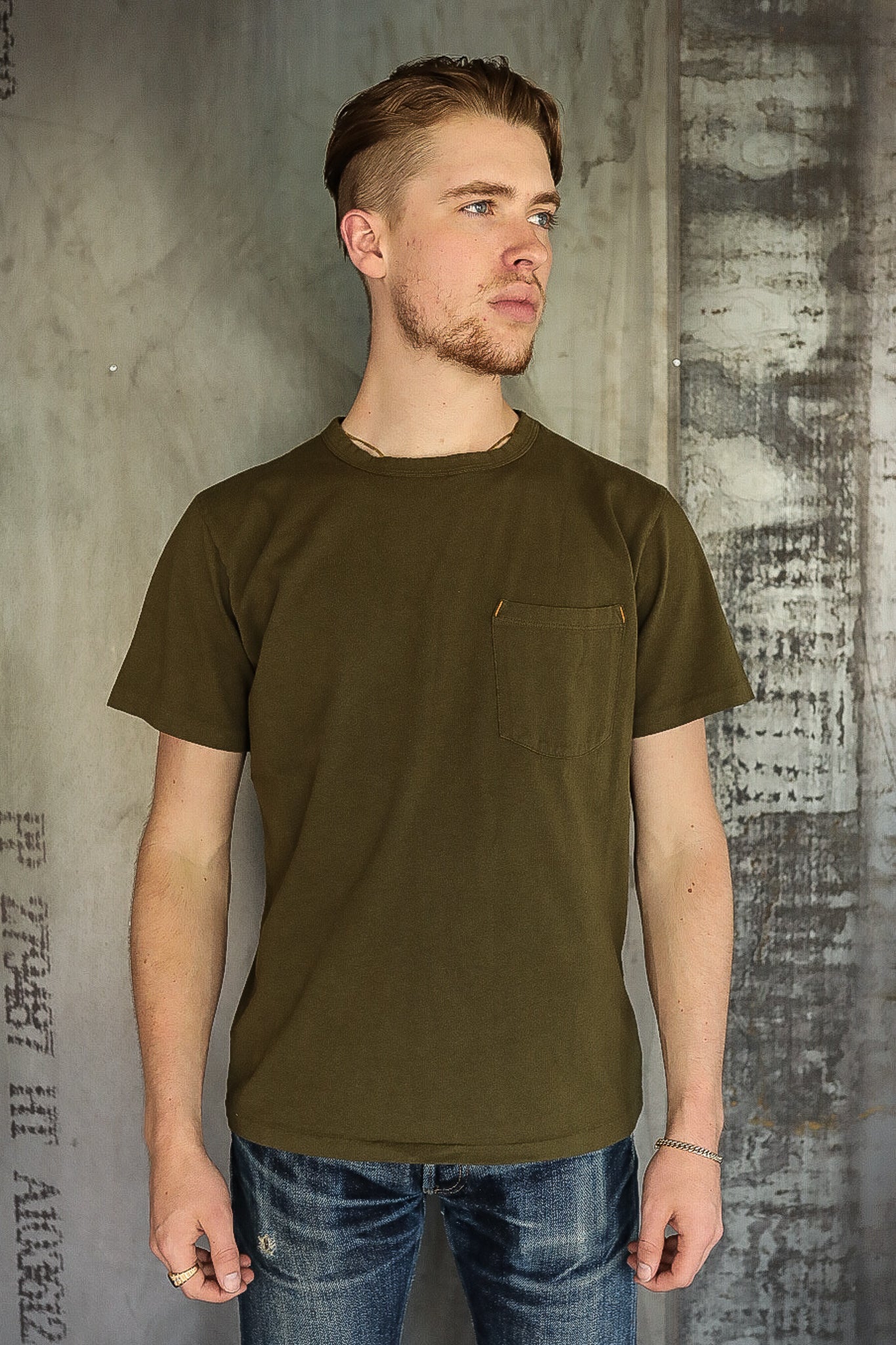 Man wearing olive green short sleeve pocket t-shirt and distressed blue jeans against textured concrete wall