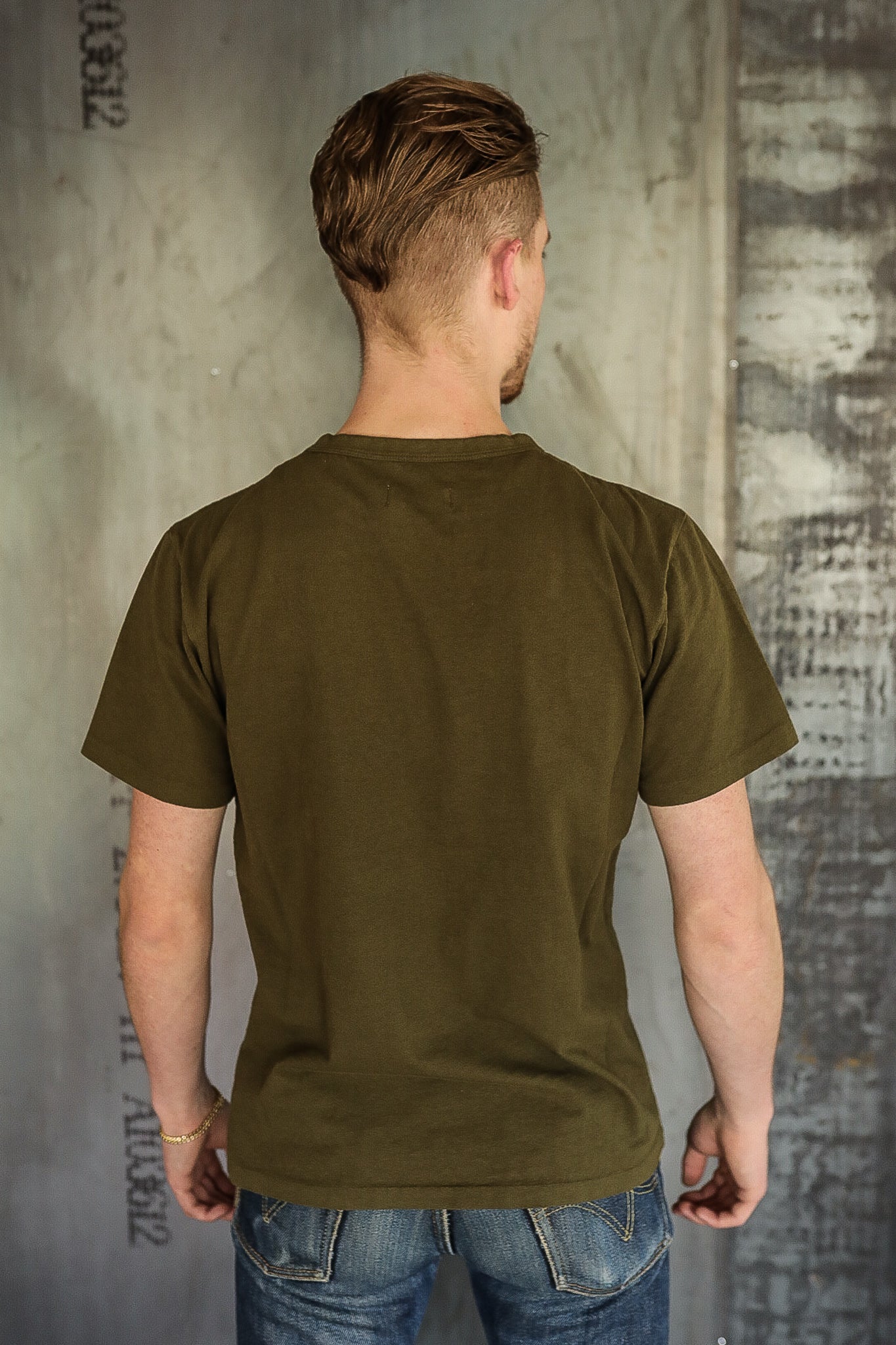 Man wearing olive green t-shirt and blue jeans standing with back to camera against textured industrial wall