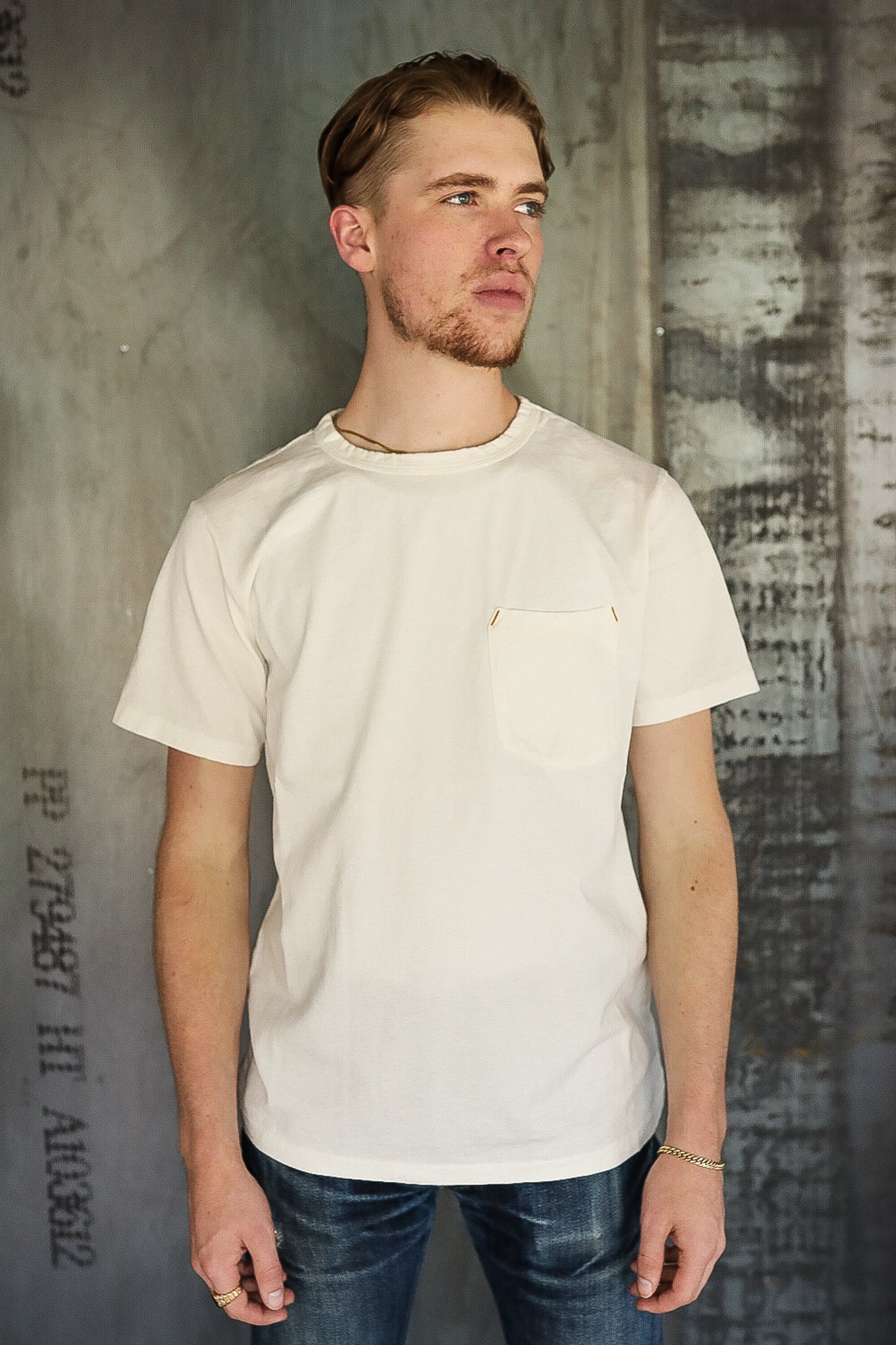 Young man wearing off-white pocket T-shirt and dark jeans against industrial textured wall