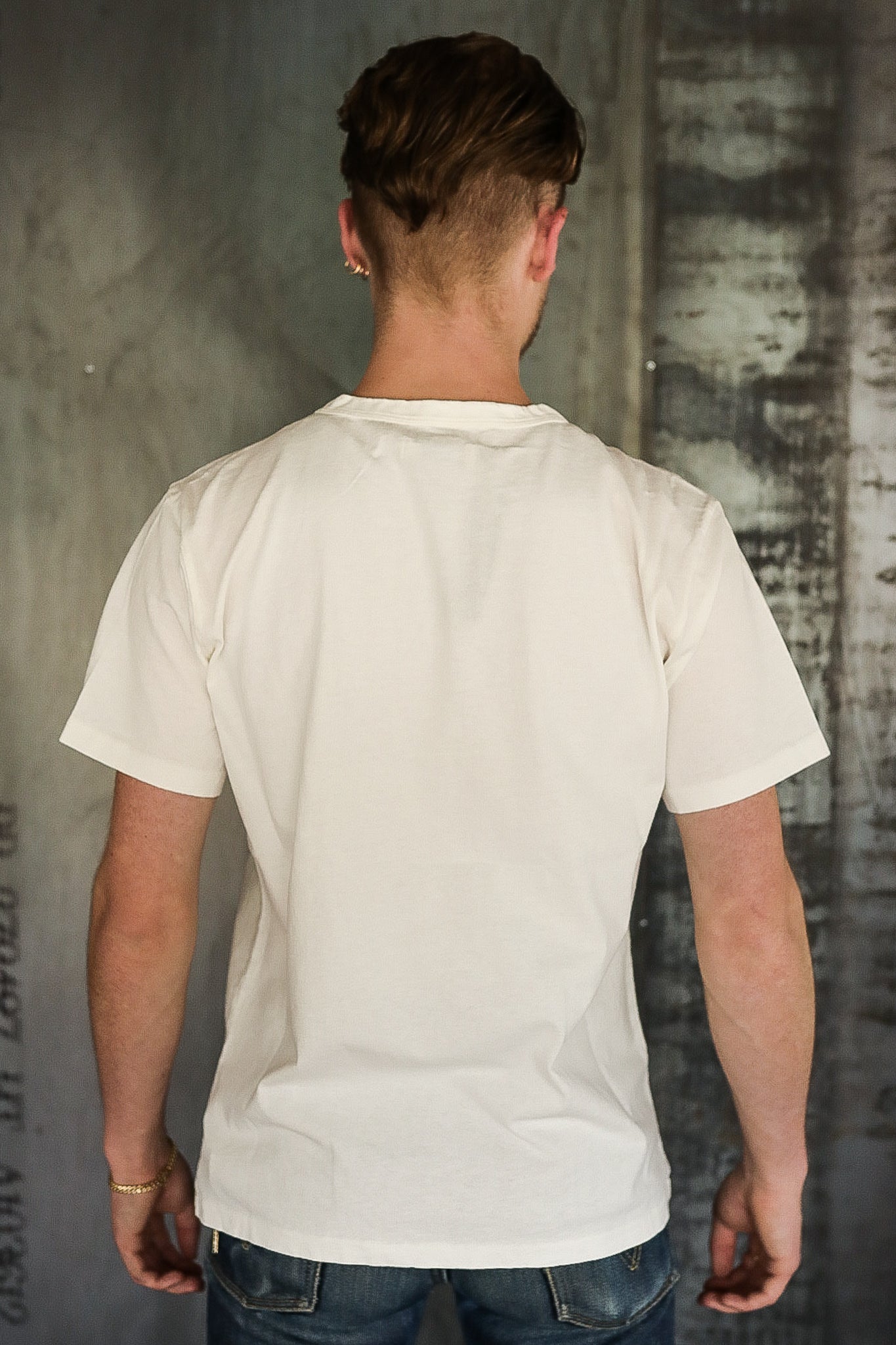 Back view of man wearing plain off-white t-shirt and blue jeans against industrial wall