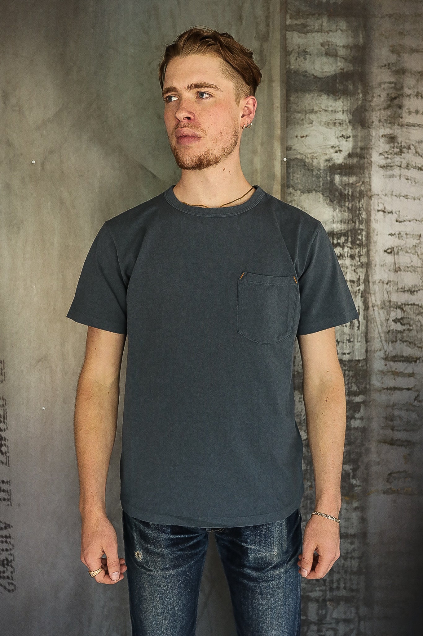 Man wearing dark gray pocket t-shirt and jeans against textured industrial wall in fashion apparel