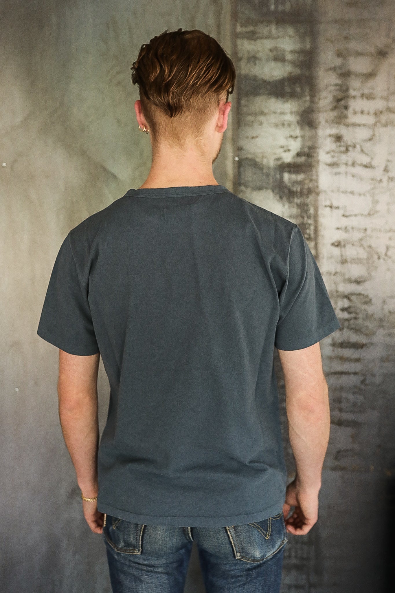 Man wearing a plain dark gray t-shirt and blue jeans standing with back to camera against textured wall