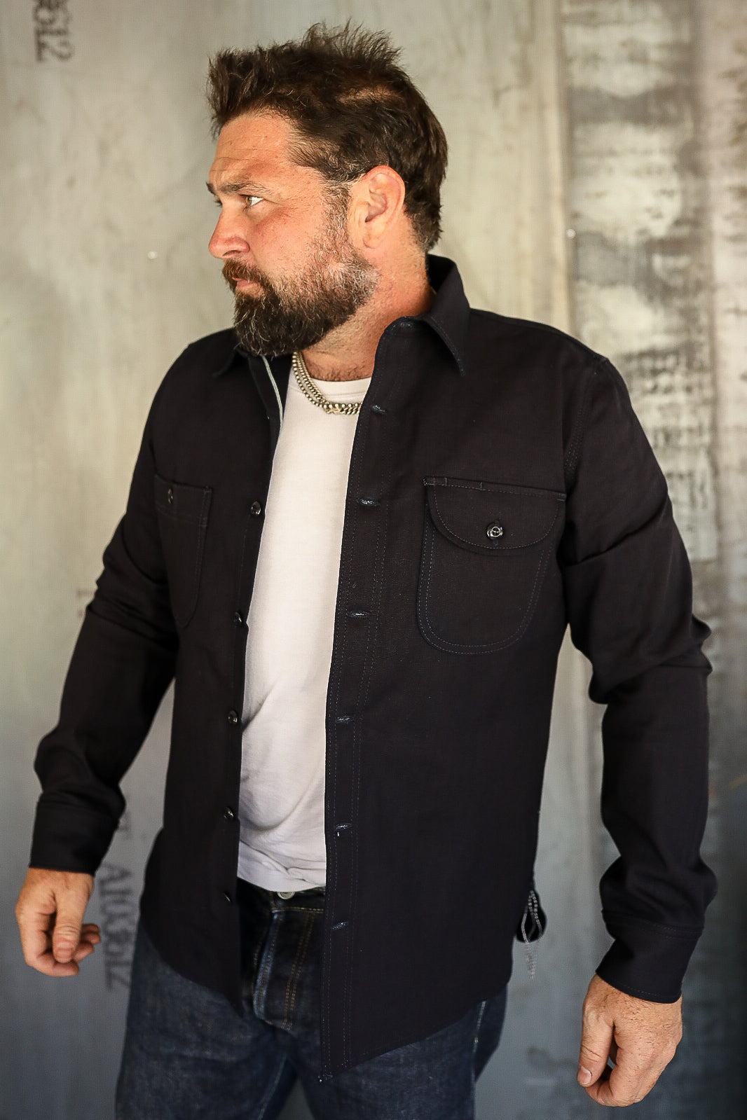 Bearded man wearing black button-up shirt over white tee, styled with chain necklace, standing against textured wall